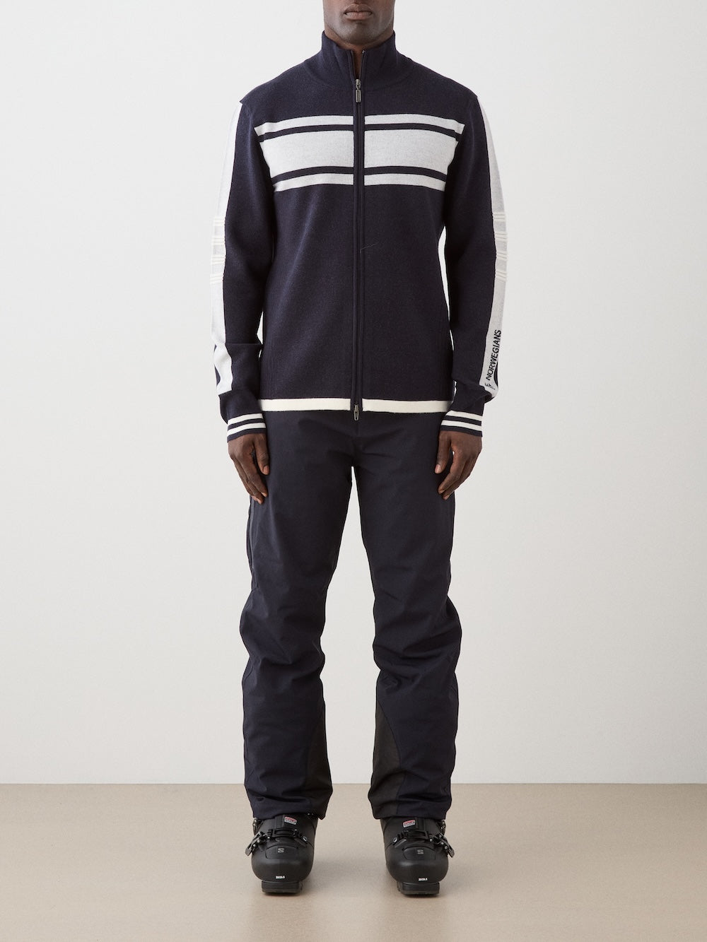 We Norwegians | FW25 | Mens Rocky Full ZipUp_Navy blue | Model wearing this fine knitted sweater with a 2-way front zipper opening, designed in a retro style with contrasting color blocks on the chest and sleeves - paired with black ski pants | Shot against an off-white background | Front