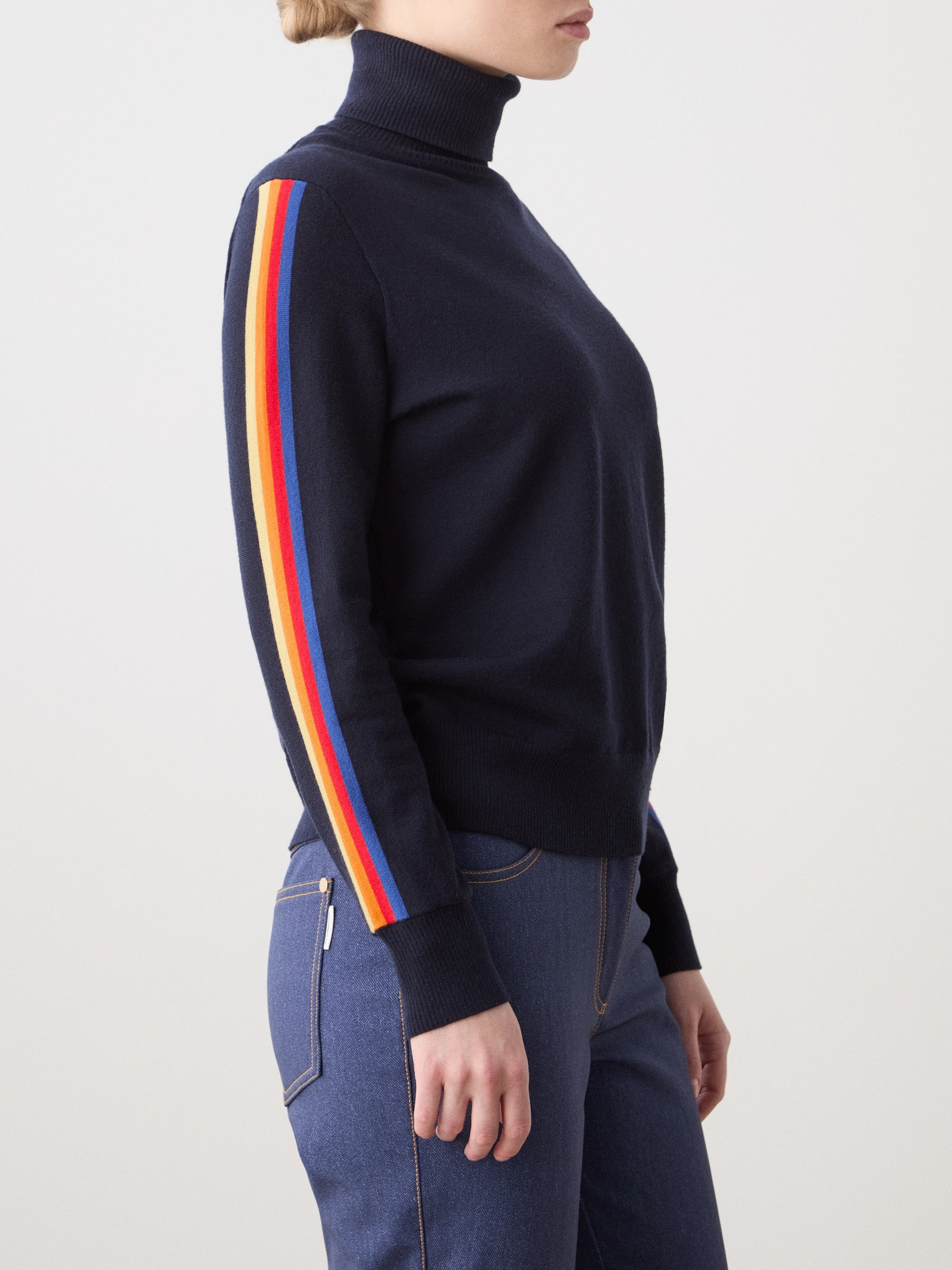 We Norwegians | FW25 | Womens Freebird Turtleneck Sweater_Navy blue | Model is wearing this knitted turtleneck sweater with contrasting panels on the sleeves - paired with the Western ski pants | Shot against an off-white background | Side/ Close-up