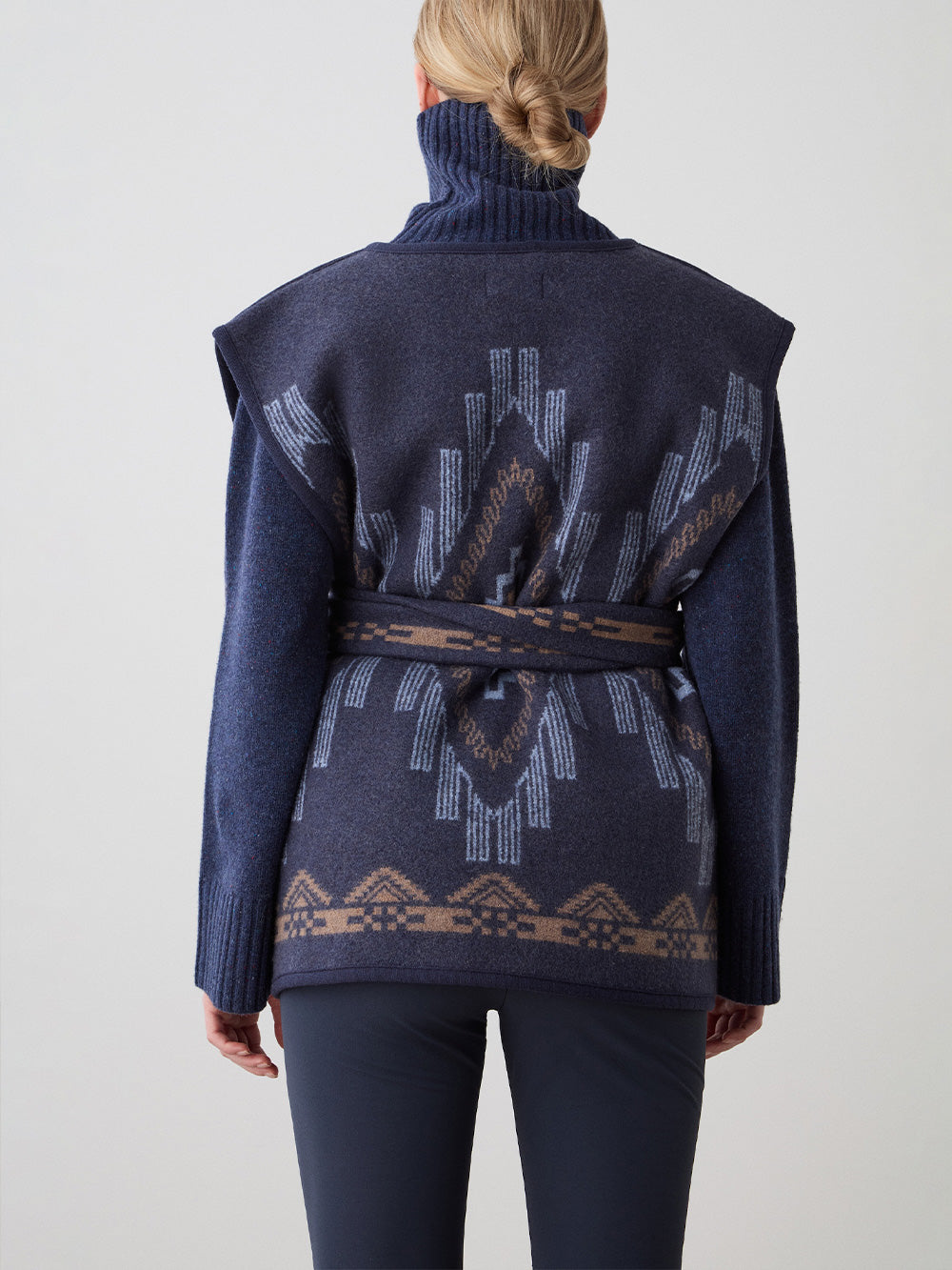 We Norwegians_FW25_Women | Model is wearing the Kautokeino vest | Navy blue in color with a jacquard pattern inspired by Native and Indigenous American Art | Vest has a matching belt to cinch the waist | Back view on a white background