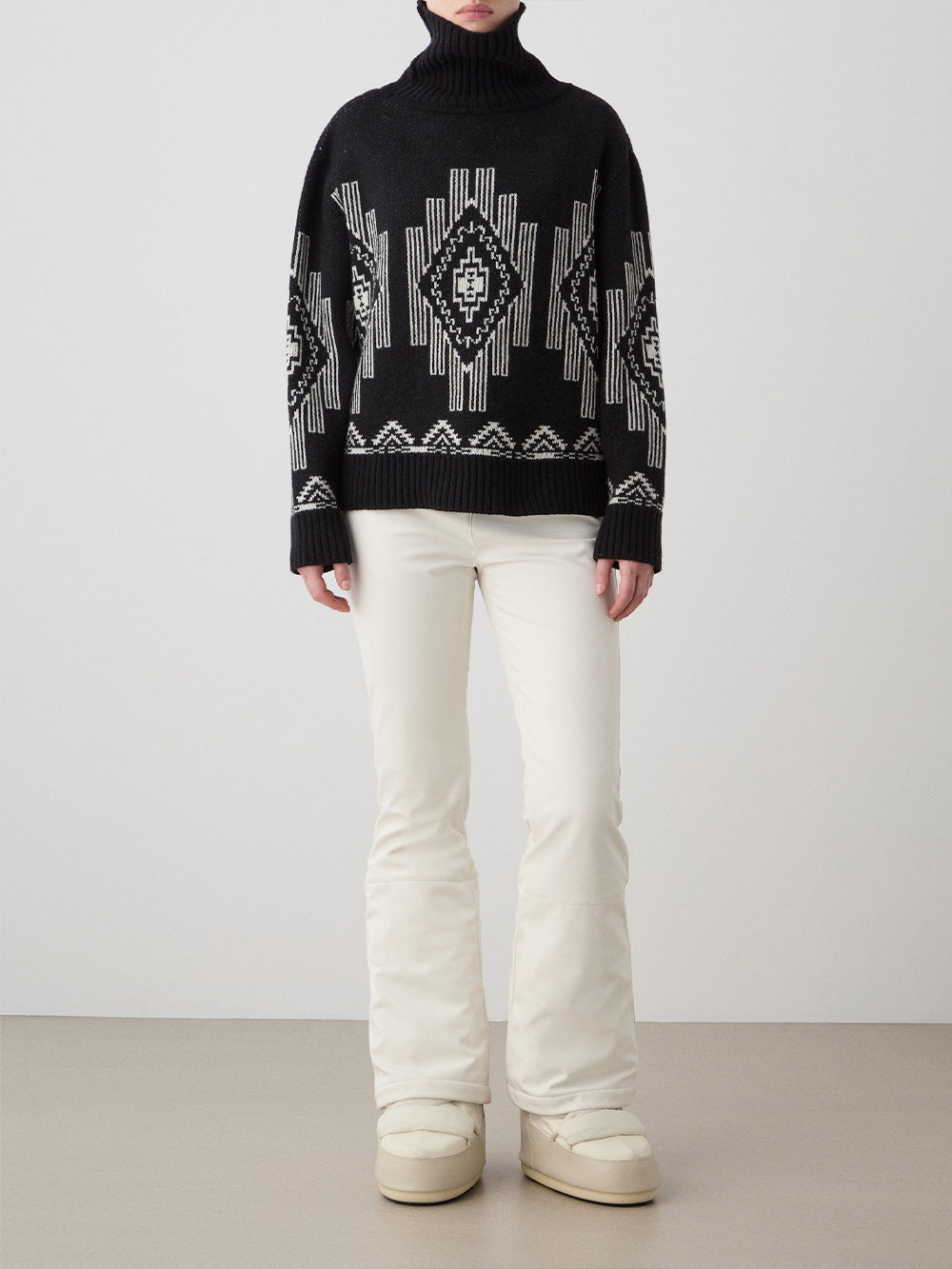 A woman wears the We Norwegians Kautokeino Sweater—black wool with a white geometric pattern—paired with off-white pants and boots, standing against a plain, light background.