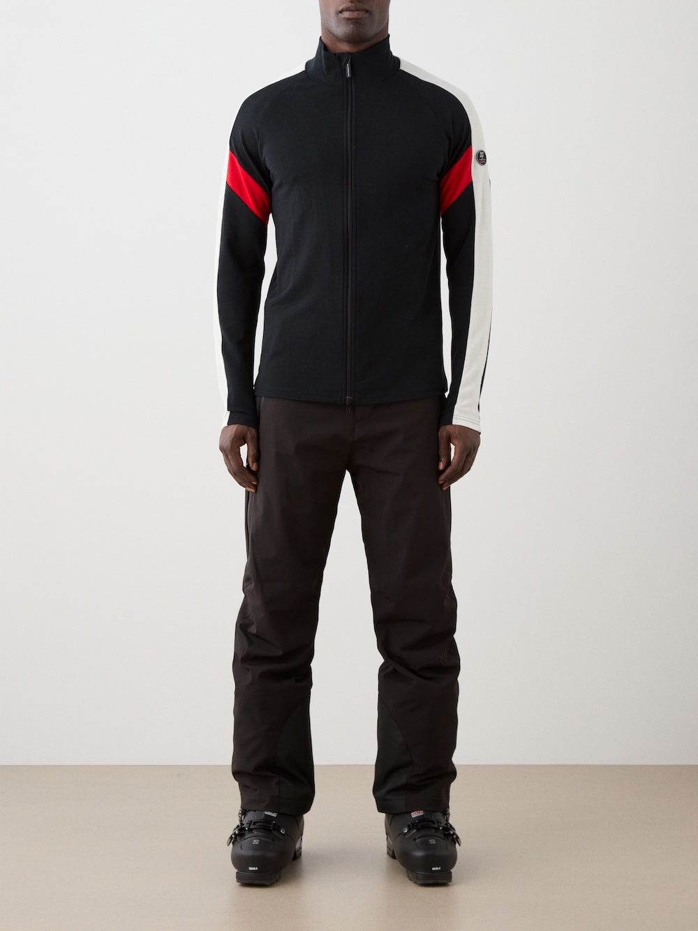 We Norwegians | FW25 | Mens Zermatt Full Zip Sweater_Black | The model is wearing this piqué knit sweater with white and red accents on the sleeves and a front 2-way zipper opening - paired with black ski pants | Shot against an off-white background | Front