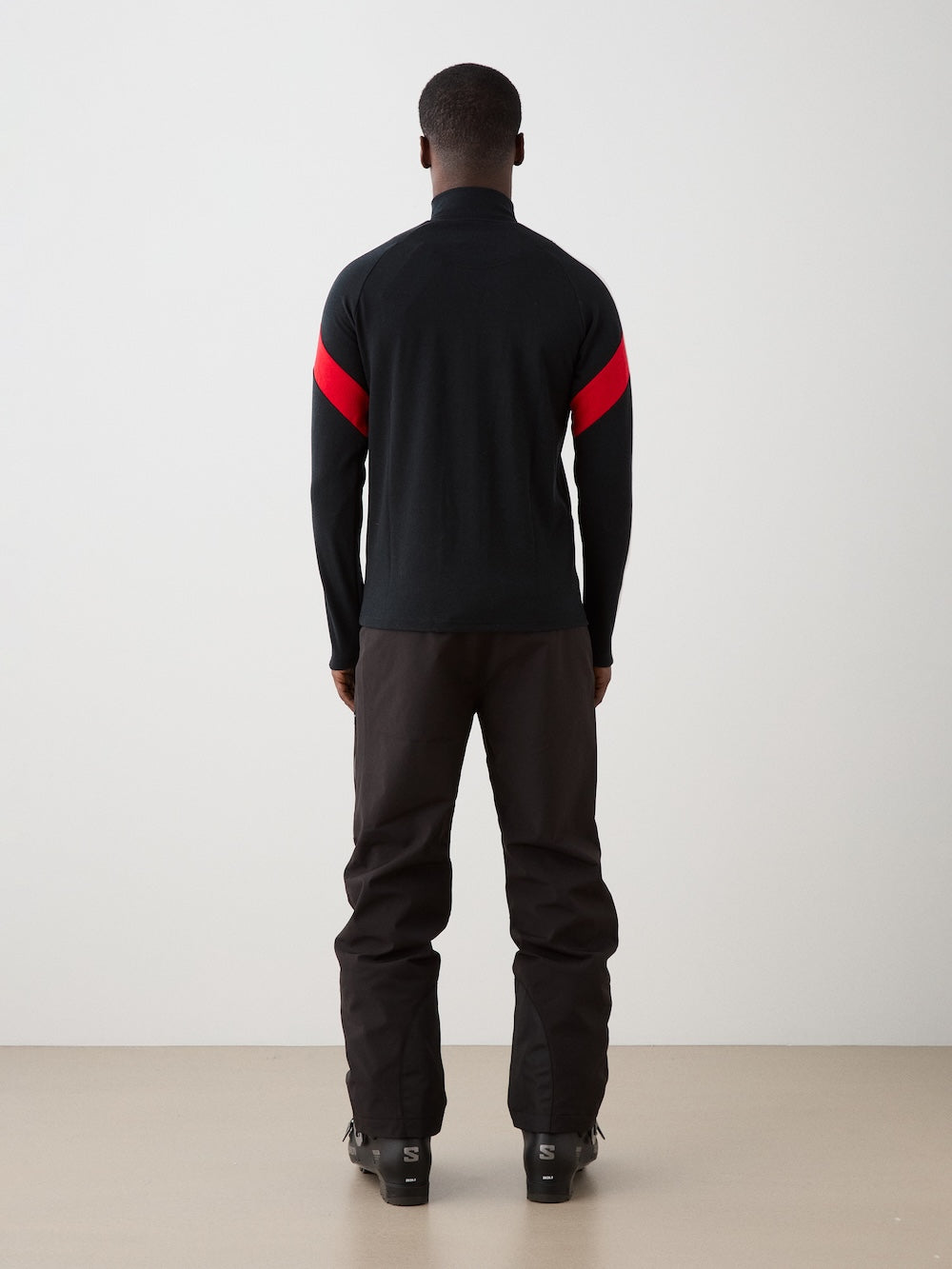 We Norwegians | FW25 | Mens Zermatt Full Zip Sweater_Black | The model is wearing this piqué knit sweater with white and red accents on the sleeves and a front 2-way zipper opening - paired with black ski pants | Shot against an off-white background | Back