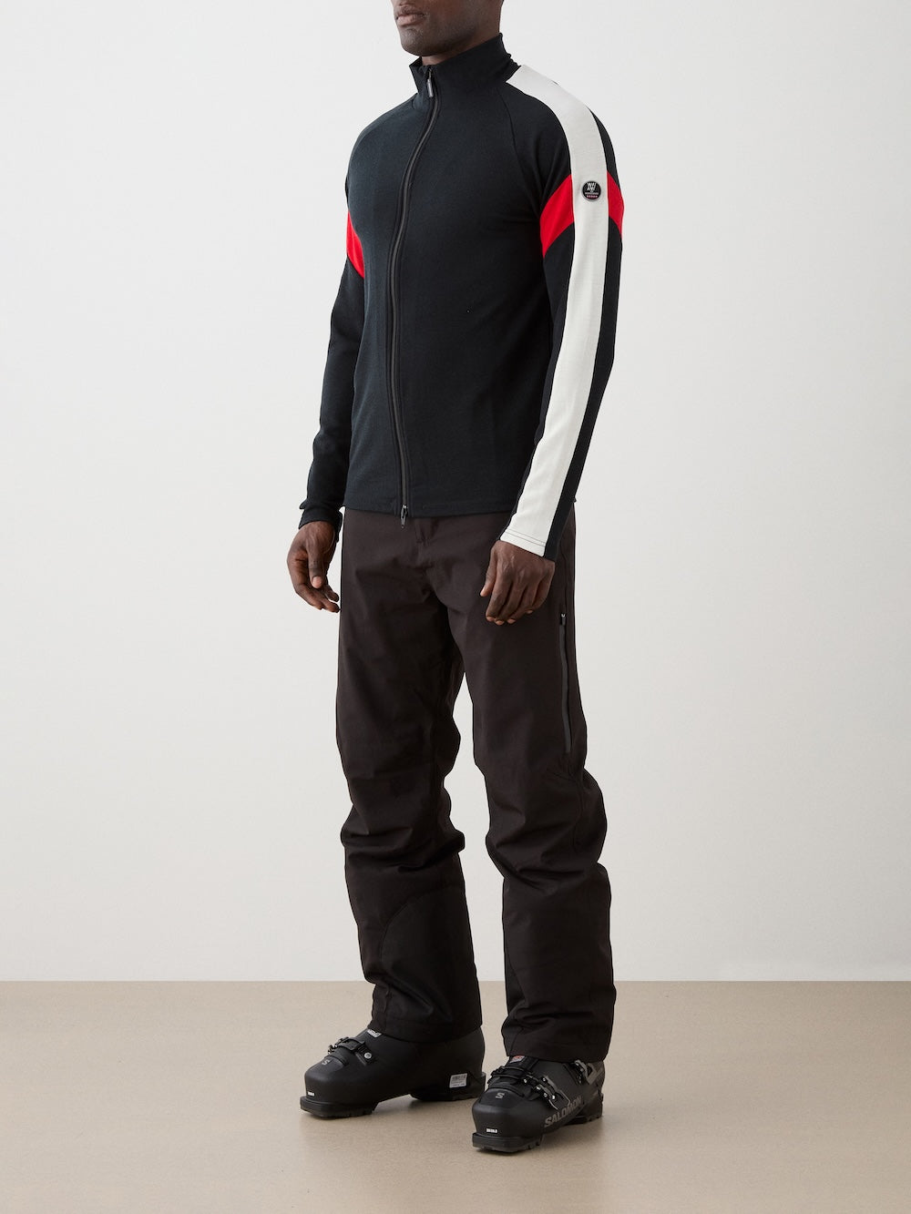 We Norwegians | FW25 | Mens Zermatt Full Zip Sweater_Black | The model is wearing this piqué knit sweater with white and red accents on the sleeves and a front 2-way zipper opening - paired with black ski pants | Shot against an off-white background | Side