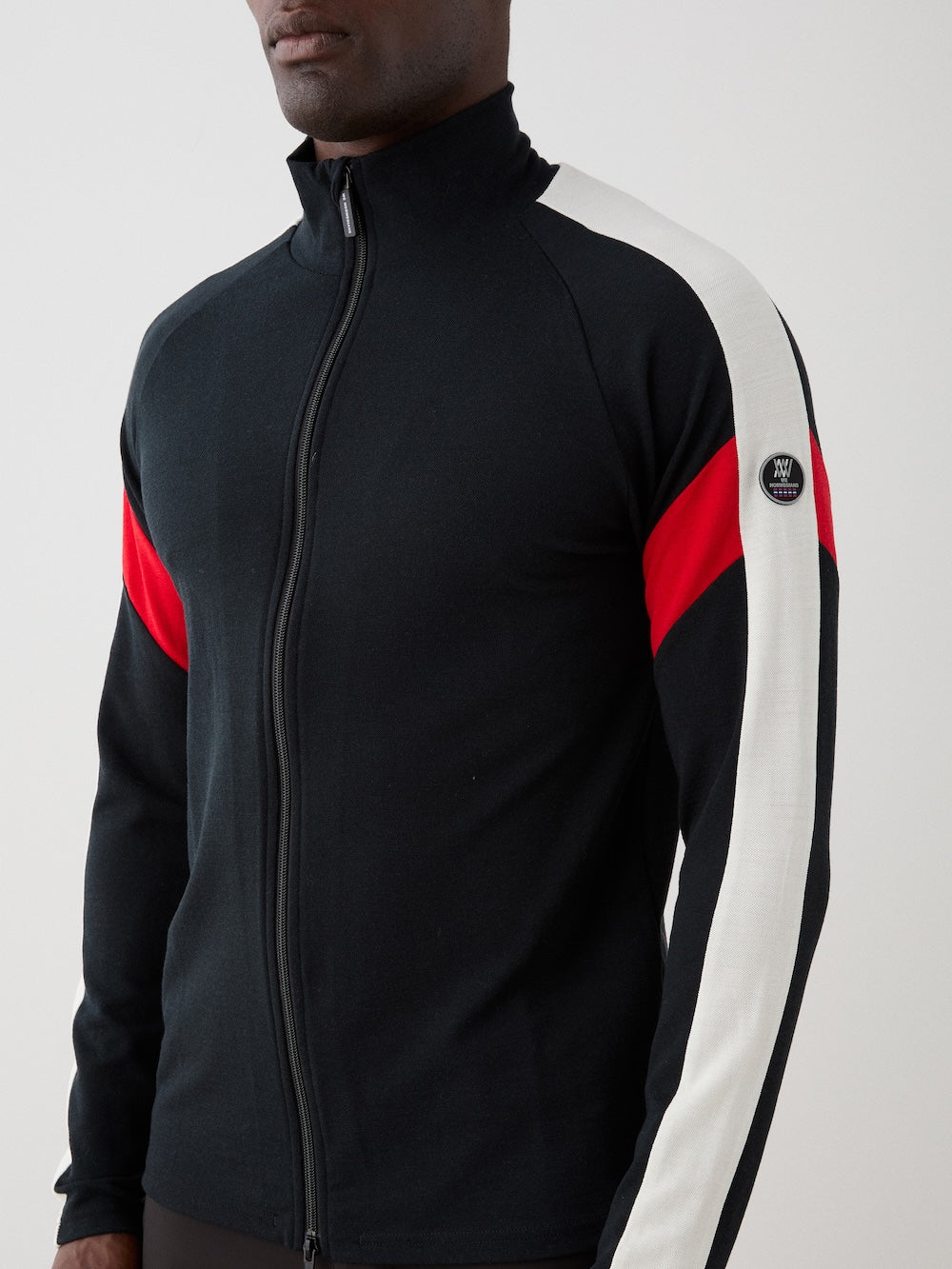 We Norwegians | FW25 | Mens Zermatt Full Zip Sweater_Black | The model is wearing this piqué knit sweater with white and red accents on the sleeves and a front 2-way zipper opening | Shot against an off-white background | Close-up