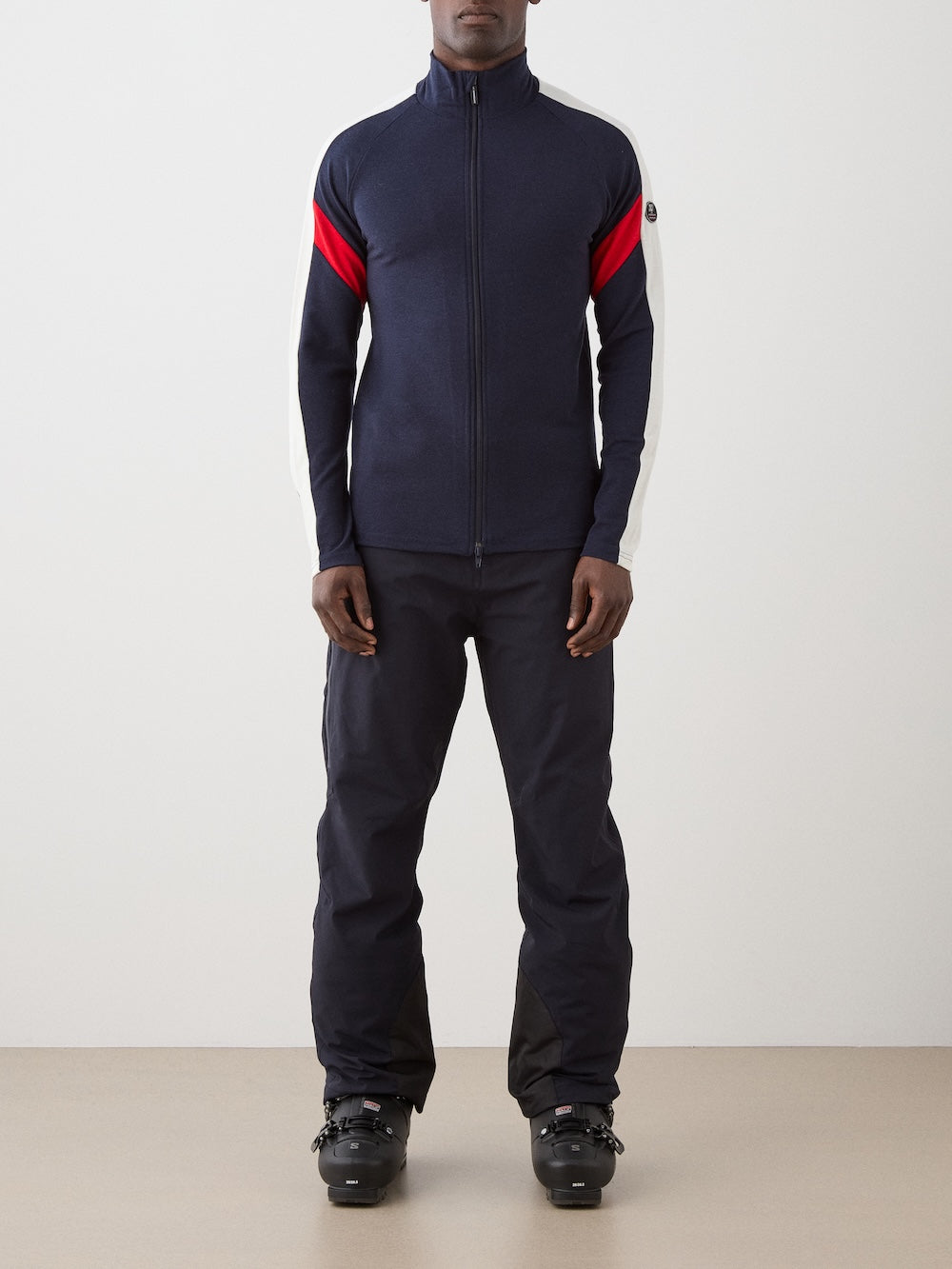 We Norwegians | FW25 | Mens Zermatt Full Zip Sweater_Flag | The model is wearing this piqué knit sweater with white and red accents on the sleeves and a front 2-way zipper opening - paired with black ski pants | Shot against an off-white background | Front