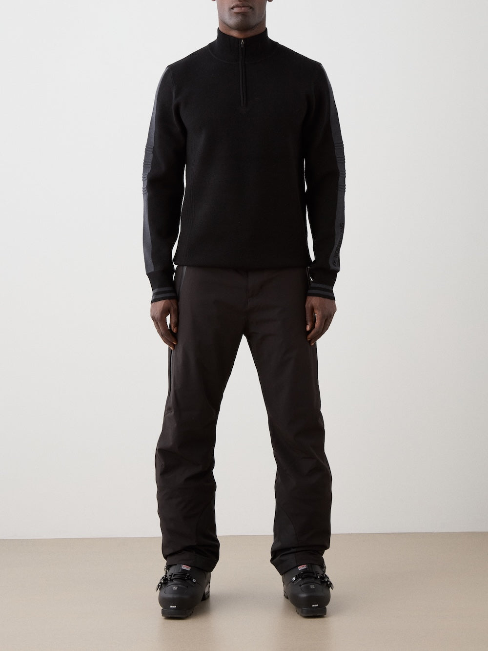 We Norwegians | FW25 | Mens Rocky ZipUp_Black | The model is wearing this knitted sweater designed in a retro style with a front 1/4th zipper opening, color block panels on the sleeves, with embroidered branding - paired with black ski pants | Front