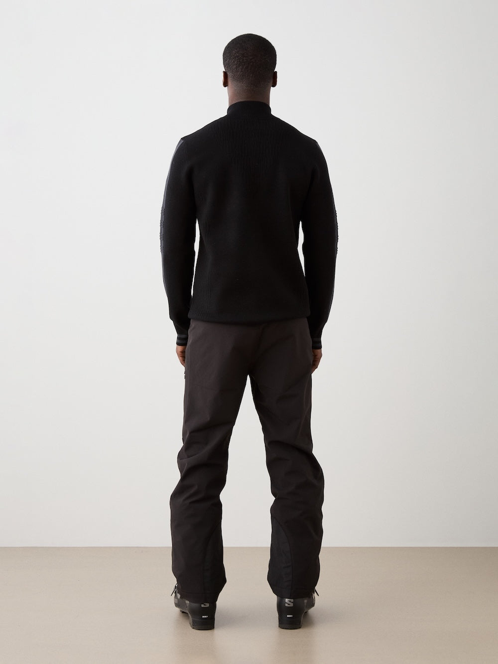 We Norwegians | FW25 | Mens Rocky ZipUp_Black | The model is wearing this knitted sweater designed in a retro style with a front 1/4th zipper opening, color block panels on the sleeves, with embroidered branding - paired with black ski pants | Back