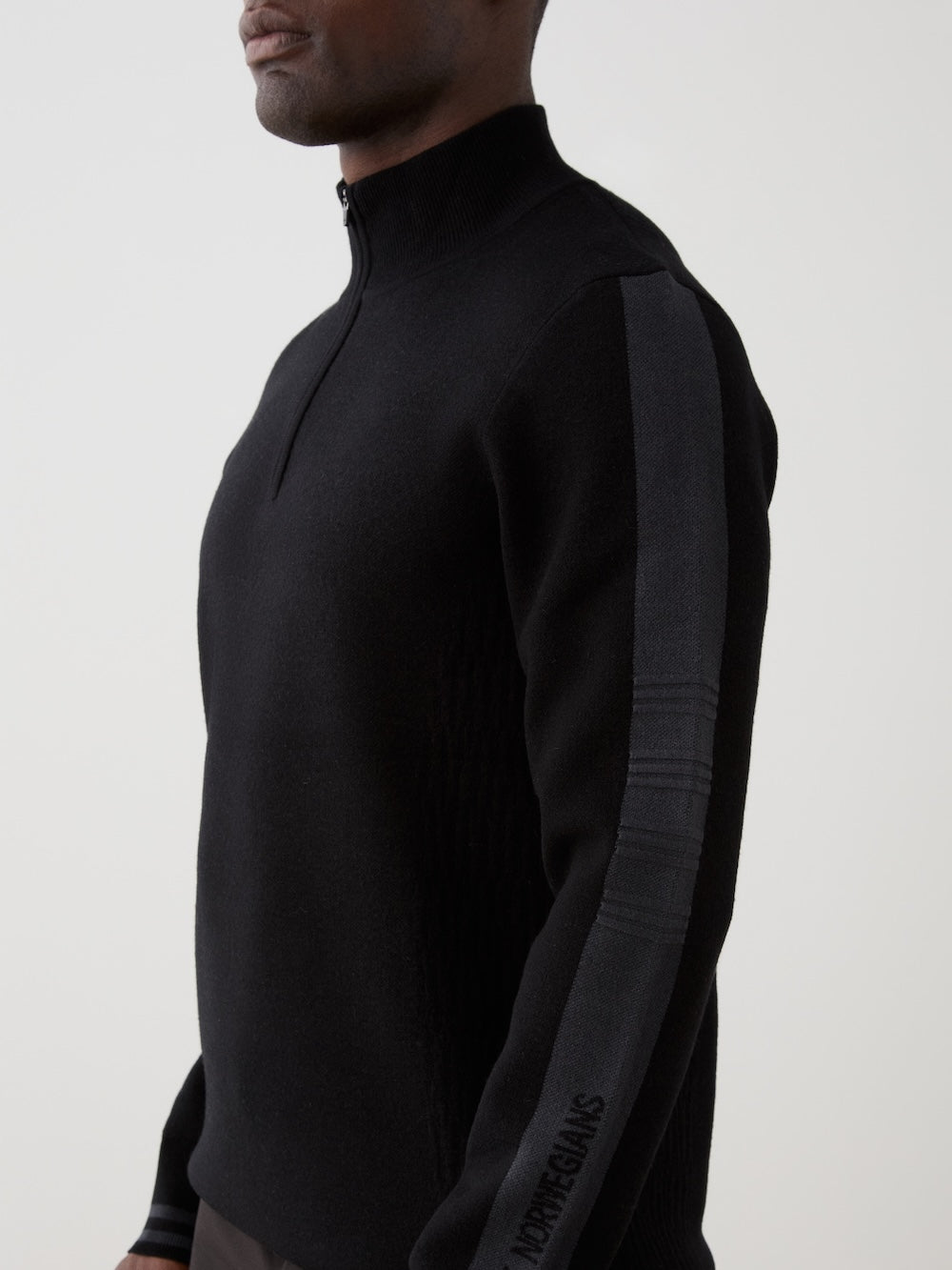 We Norwegians | FW25 | Mens Rocky ZipUp_Black | The model is wearing this knitted sweater designed in a retro style with a front 1/4th zipper opening, color block panels on the sleeves, with embroidered branding | Close-up