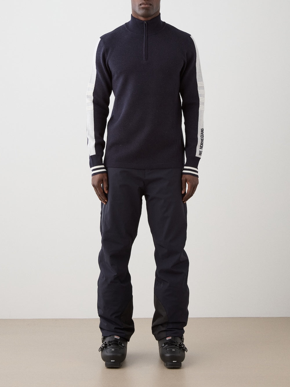 We Norwegians | FW25 | Mens Rocky ZipUp_Navy blue | The model is wearing this knitted sweater designed in a retro style with a front 1/4th zipper opening, color block panels on the sleeves, with embroidered branding - paired with black ski pants | Front