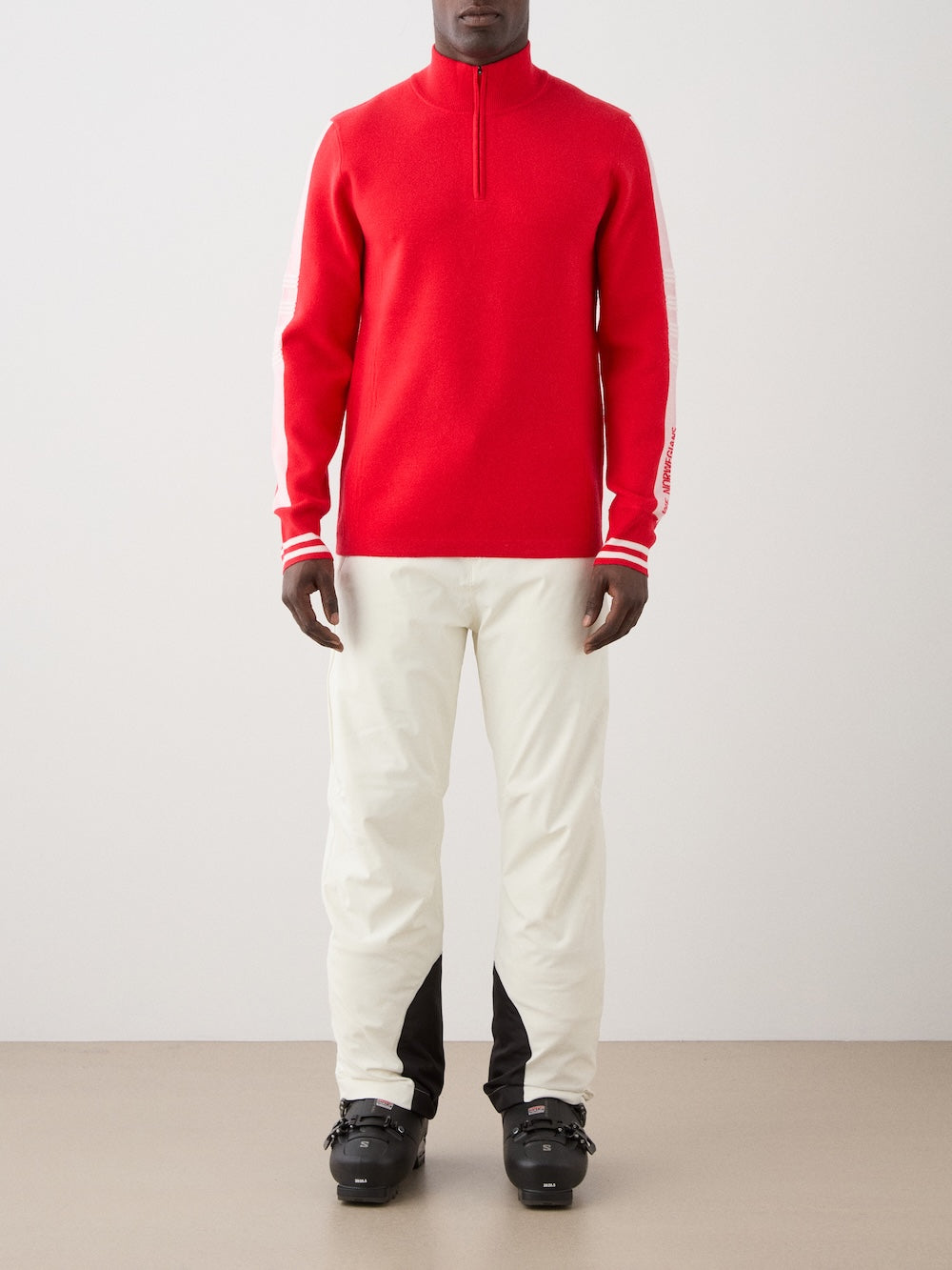 We Norwegians | FW25 | Mens Rocky ZipUp_Red | The model is wearing this knitted sweater designed in a retro style with a front 1/4th zipper opening, color block panels on the sleeves, with embroidered branding - paired with white ski pants | Front