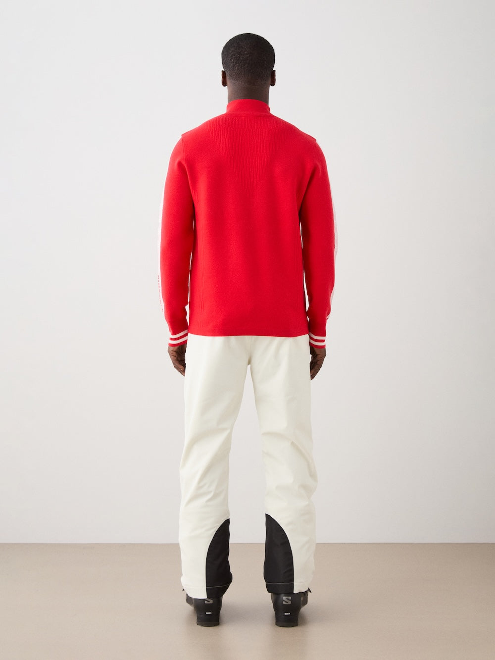 We Norwegians | FW25 | Mens Rocky ZipUp_Red | The model is wearing this knitted sweater designed in a retro style with a front 1/4th zipper opening, color block panels on the sleeves, with embroidered branding - paired with white ski pants | Back