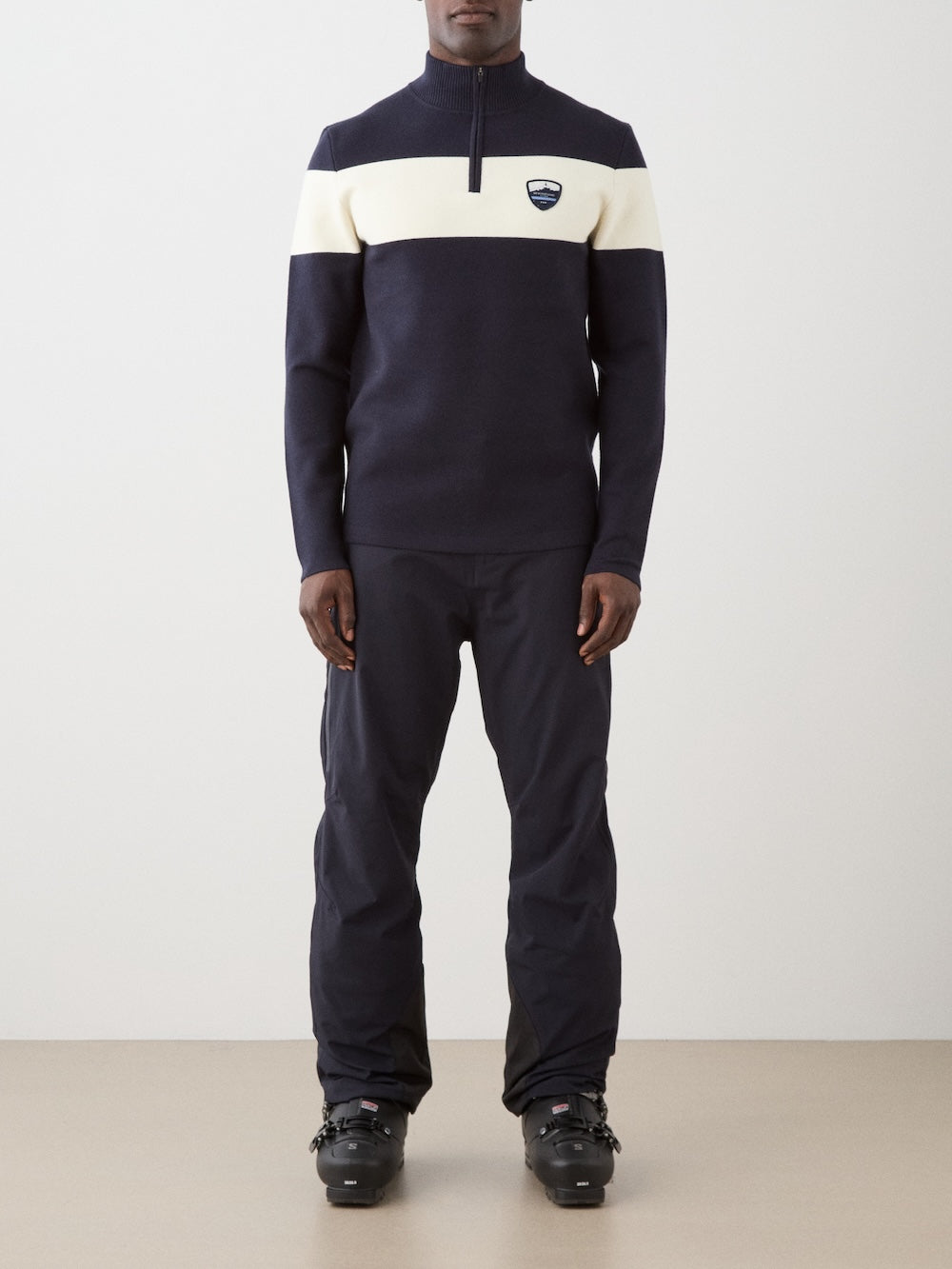 We Norwegians | FW25 | Mens Holmenkollen ZipUp_Navy blue | Model is wearing a merino/ cotton blend - knitted sweater with a 1/4th zipper opening and badge at the front - paired with black ski pants | Shot against an off-white background | Front view