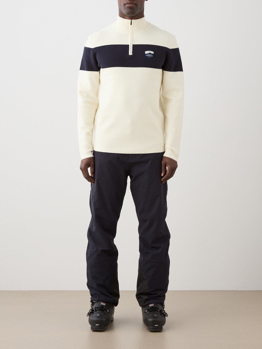 We Norwegians | FW25 | Mens Holmenkollen ZipUp_Off white | Model is wearing a merino/ cotton blend - knitted sweater with a 1/4th zipper opening and badge at the front - paired with black ski pants | Shot against an off-white background | Front view