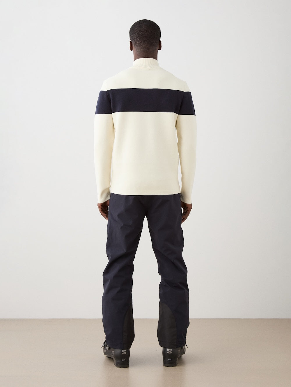 We Norwegians | FW25 | Mens Holmenkollen ZipUp_Off white | Model is wearing a merino/ cotton blend - knitted sweater with a 1/4th zipper opening and badge at the front - paired with black ski pants | Shot against an off-white background | Back view