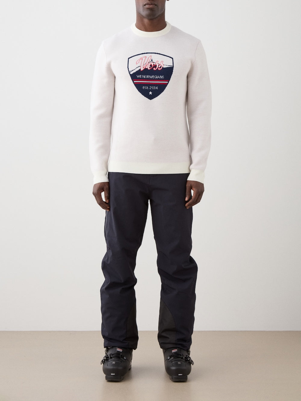 We Norwegians | FW25 | Mens Afterski Crewneck_Cream | Model wearing a knitted sweater with a design and logo at the front with black ski pants | Shot against an off-white background | Front view