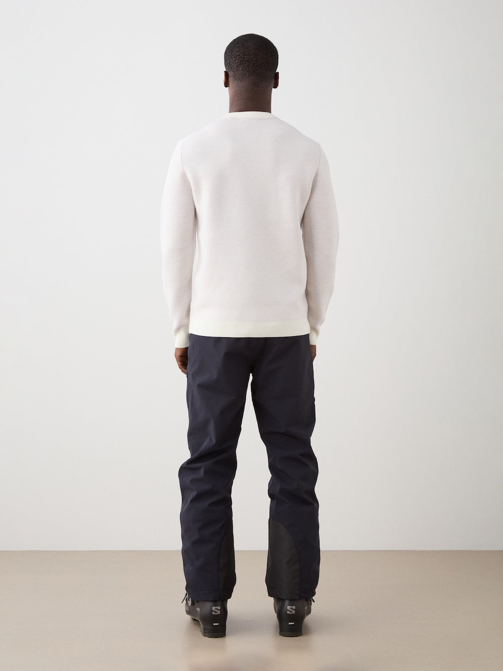 We Norwegians | FW25 | Mens Afterski Crewneck_Cream | Model wearing a knitted sweater with a design and logo at the front with black ski pants | Shot against an off-white background | Back view