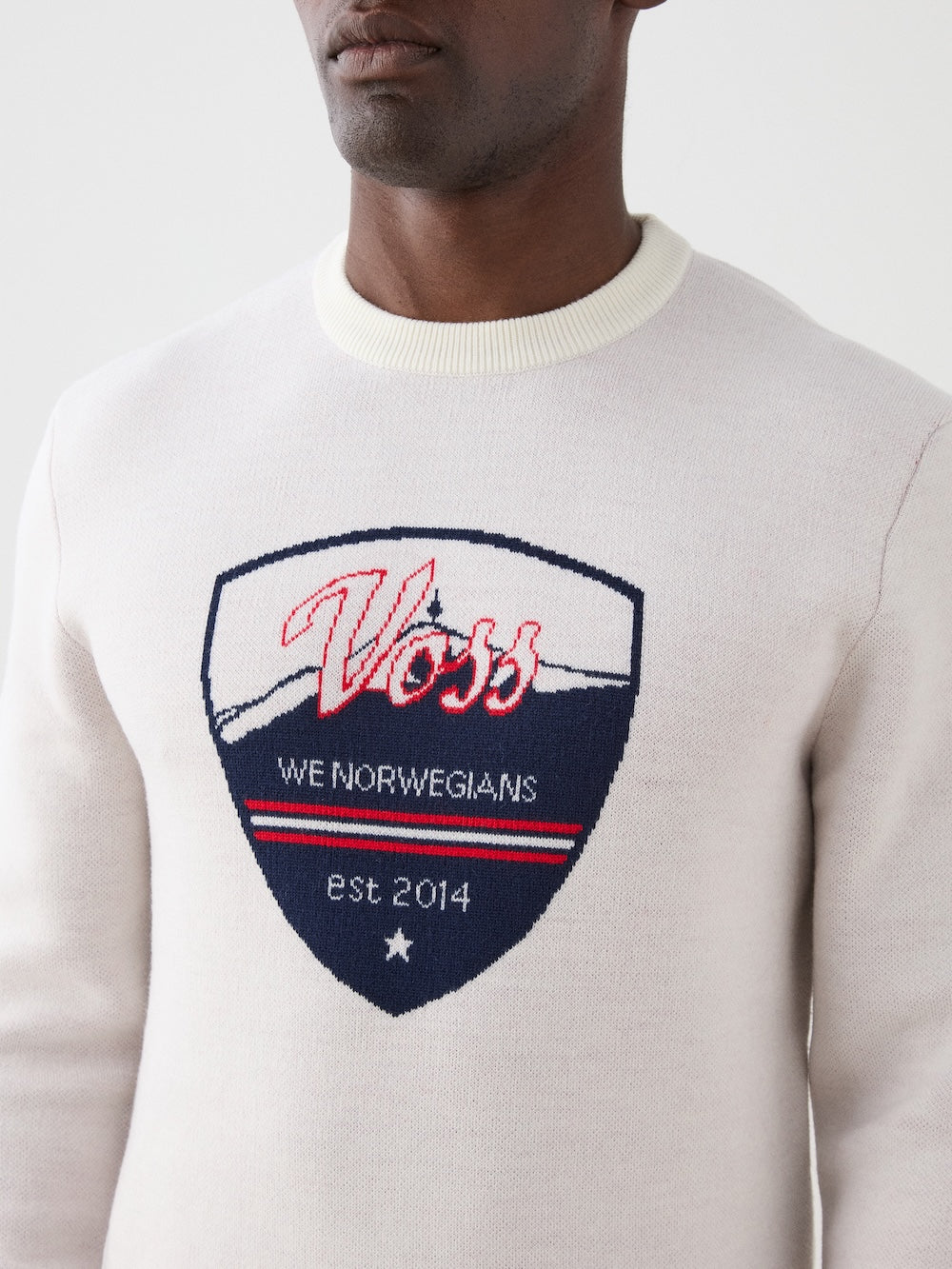 We Norwegians | FW25 | Mens Afterski Crewneck_Cream | Model wearing a knitted sweater with a design and logo at the front with black ski pants | Shot against an off-white background | Close-up view
