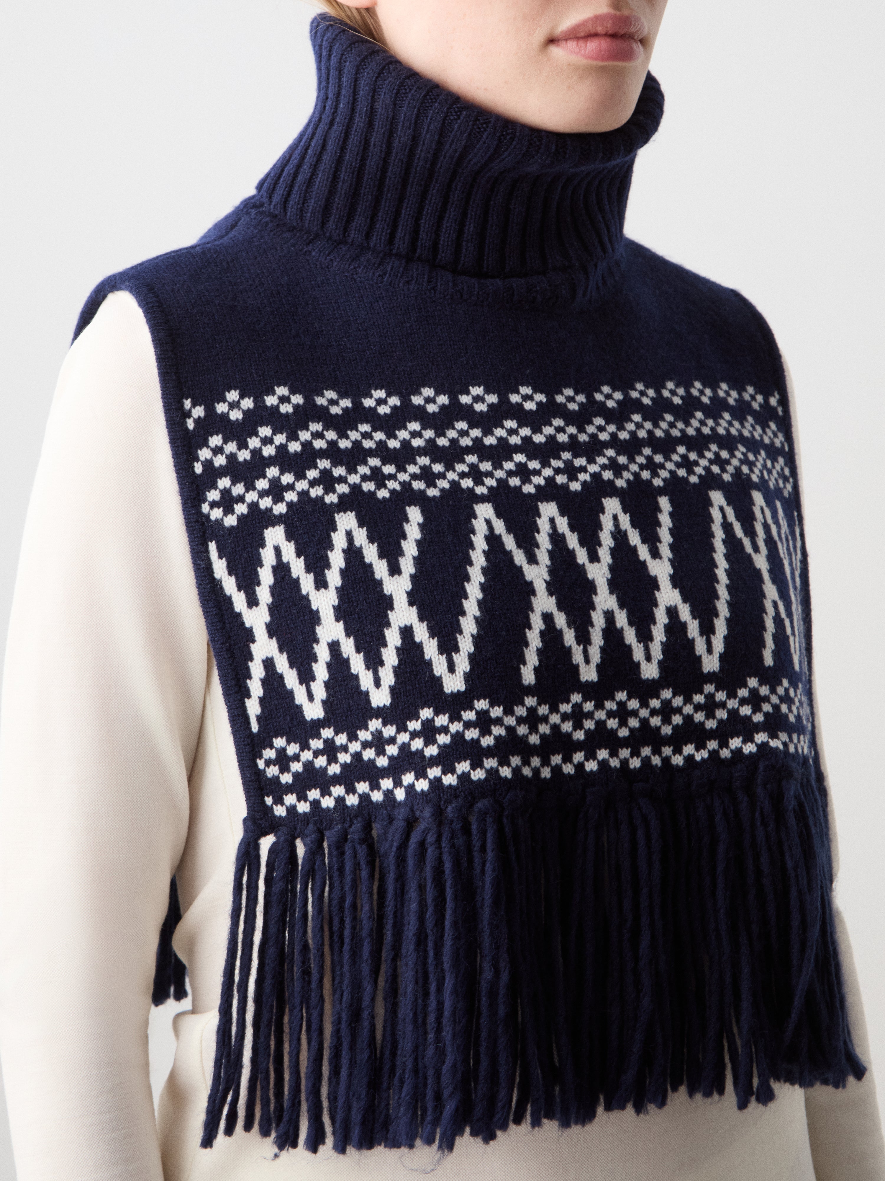 We Norwegians | FW25 | Setesdal Bib_Navy blue | Model is wearing the knitted bib with a turtle neck and fine fringes at the hem over a white sweater, against an off-white background