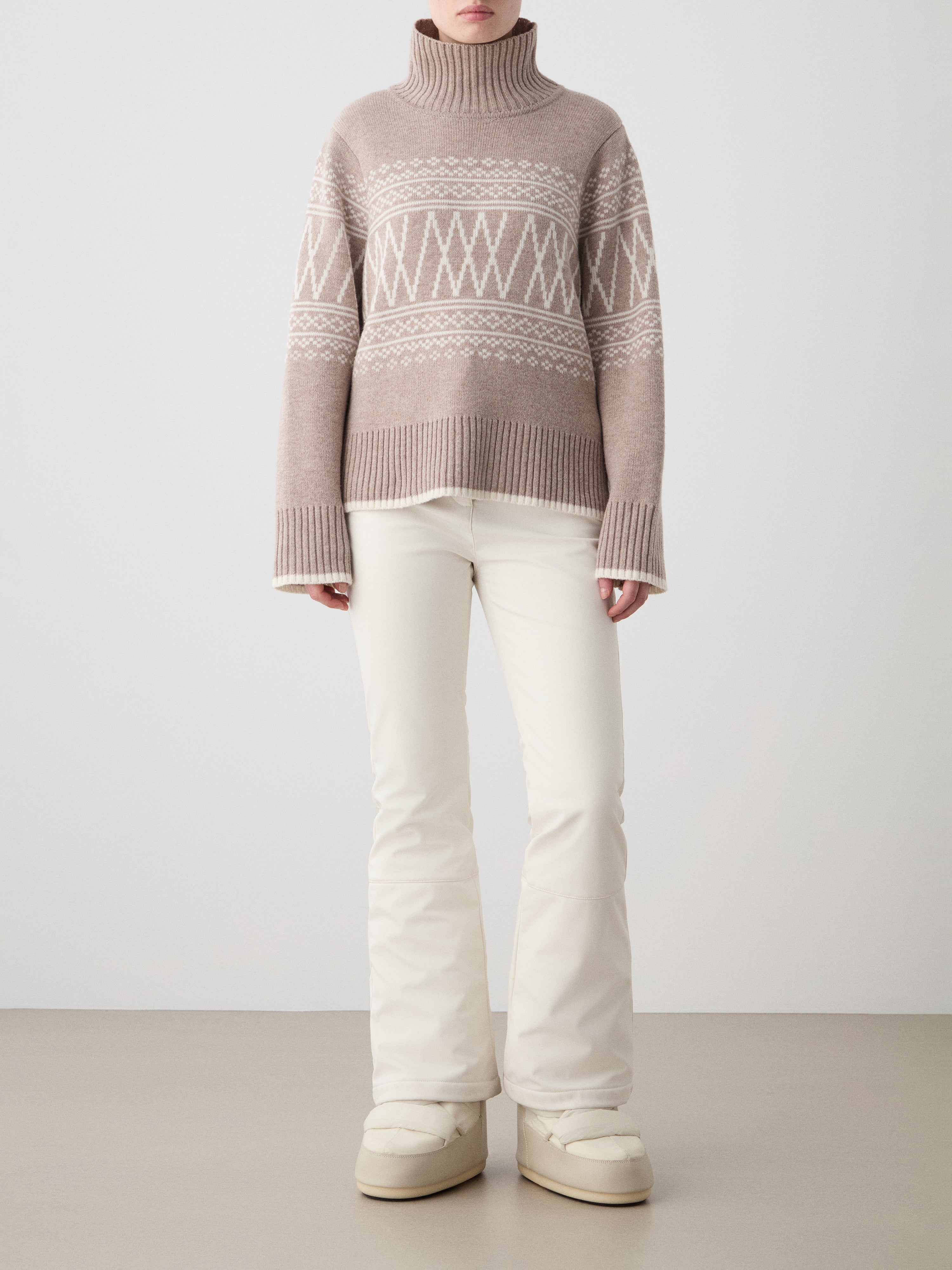 Person wearing a patterned sweater and white pants on a plain background