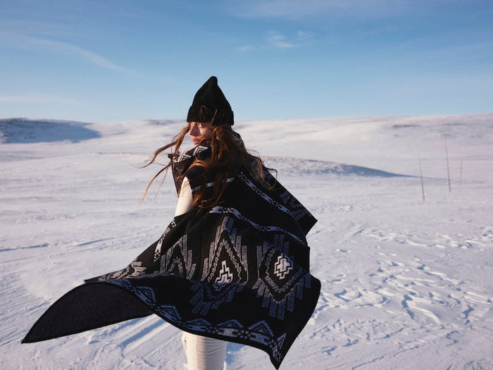 We Norwegians | FW25 | Woman wearing Kautokeino poncho in black against a backdrop of snowy Geilo