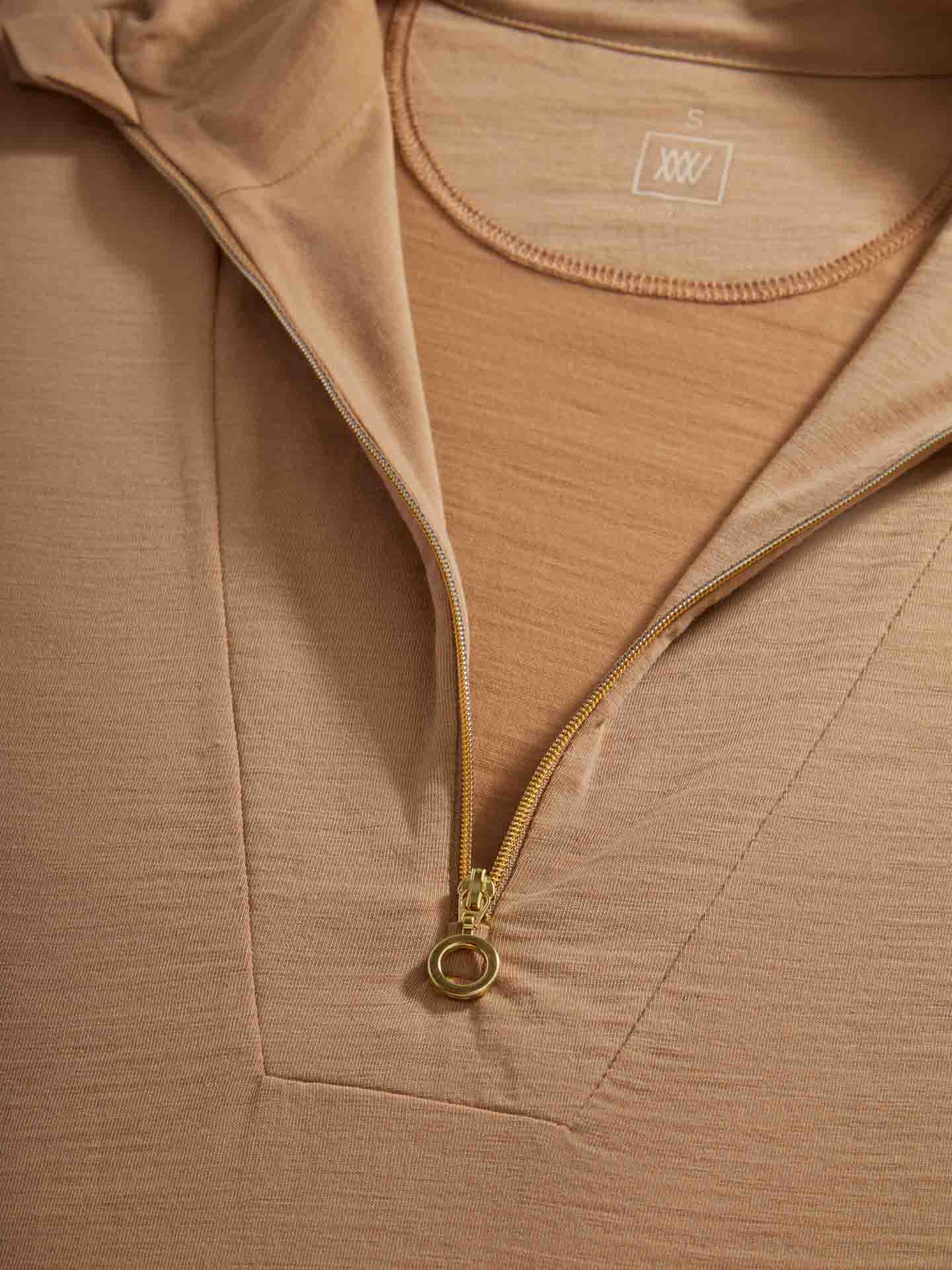 Close-up of the We Norwegians Sno Merino Zip Up Women pullover in light brown, featuring a partially unzipped gold zipper that reveals a matching inner shirt. The inside tag displays an "S" with three "X" symbols directly below it. Crafted from super fine merino wool, the fabric appears soft and finely knit, boasting a fitted shape for Scandinavian luxury base-layers.