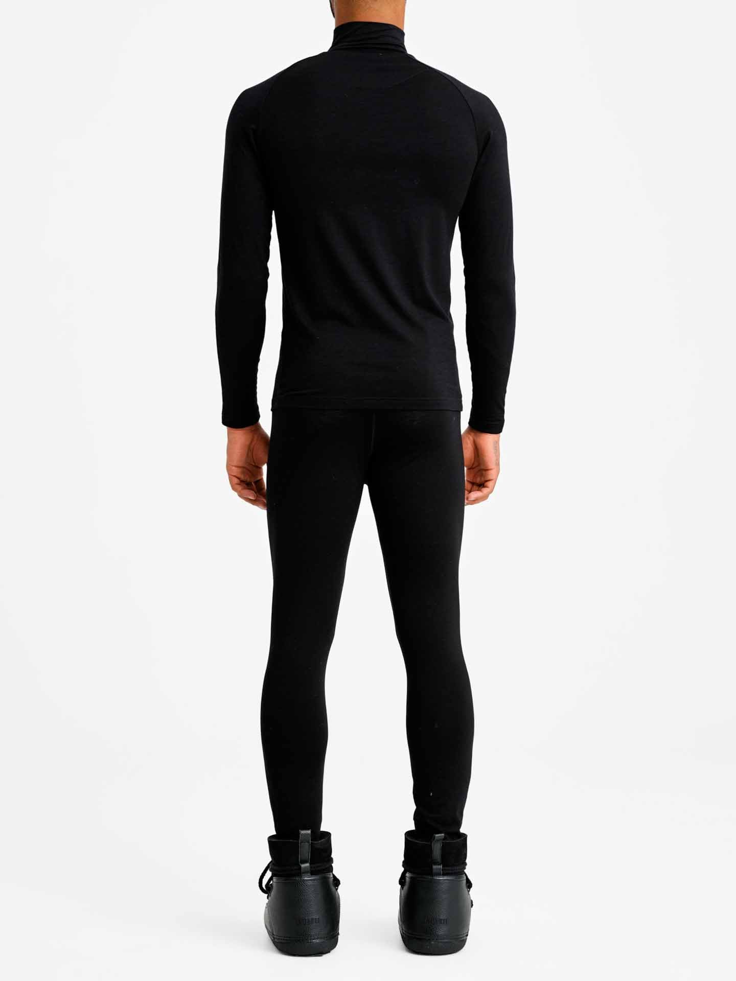 A person is standing with their back to the camera, dressed in a black We Norwegians Sno Merino Zip Up Men top, black leggings, and black boots. The background is plain white.