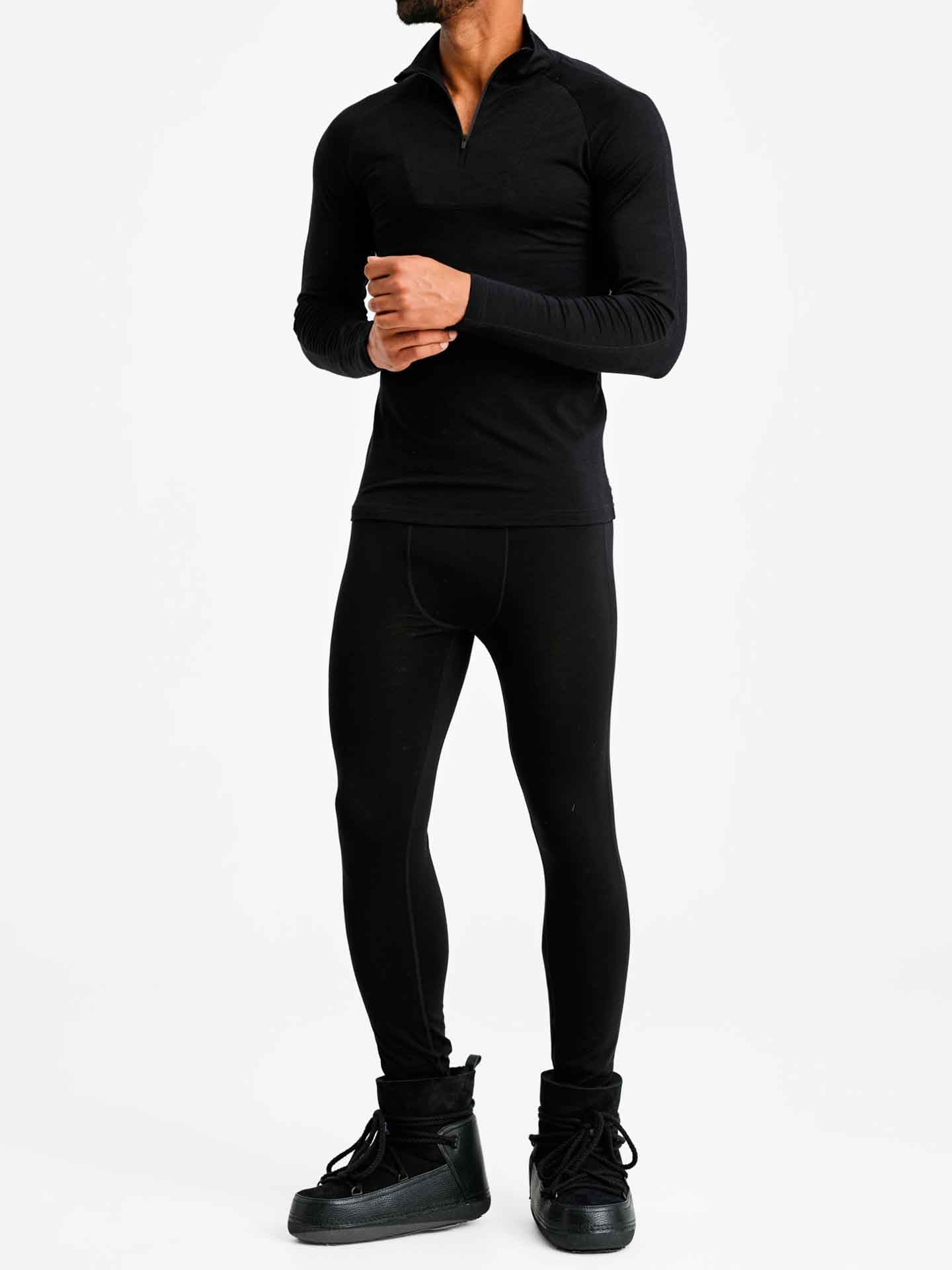 A man stands against a plain white background, dressed in black athletic wear. He is wearing the Sno Merino Zip Up Men by We Norwegians, paired with form-fitting leggings and black high-top athletic shoes. His hands are clasped together at chest level. His face is not visible, embodying Scandinavian luxury base-layers.