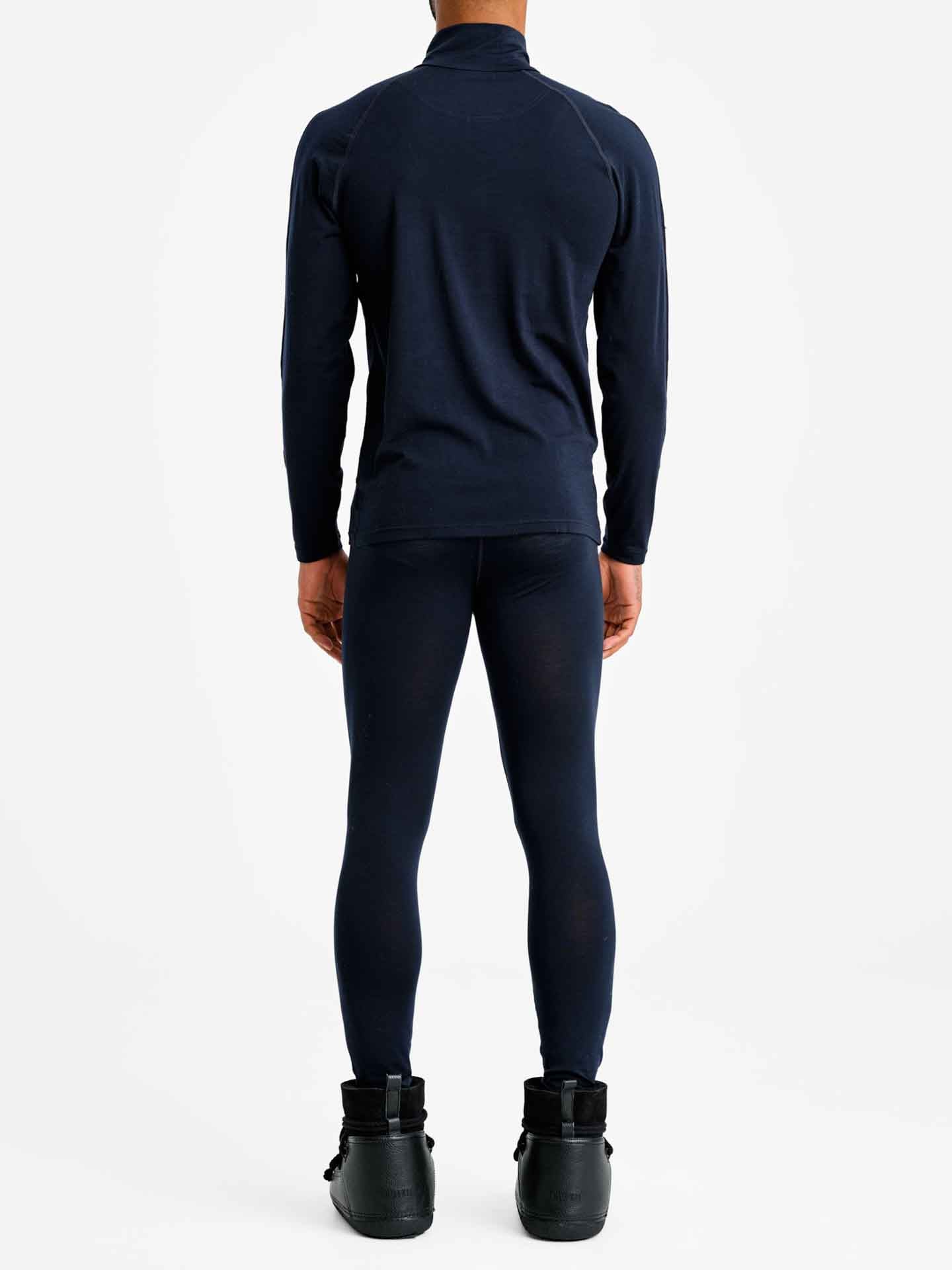 A person stands facing away from the camera, wearing the We Norwegians' Sno Merino Zip Up Men in navy paired with matching leggings made from breathable merino wool. They are also wearing black boots. The background is plain white, emphasizing the Scandinavian luxury base-layers.