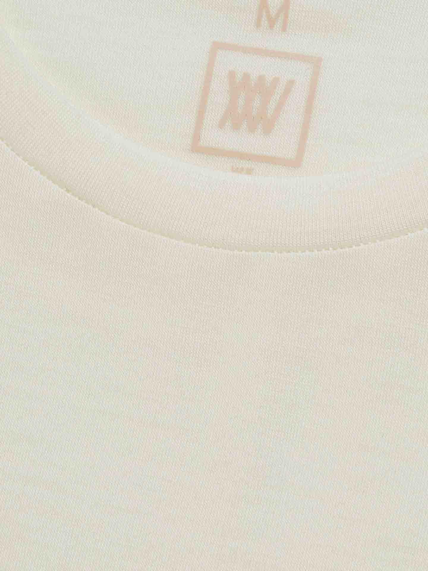 A close-up image of the Sno Merino Tee Men, a beige or off-white T-shirt made by We Norwegians from premium merino wool. The inner neck tag shows the size "M" and features a simple logo with the letters "XXIV" inside a square. The shirt fabric appears soft and finely ribbed, exemplifying Scandinavian luxury base-layers.