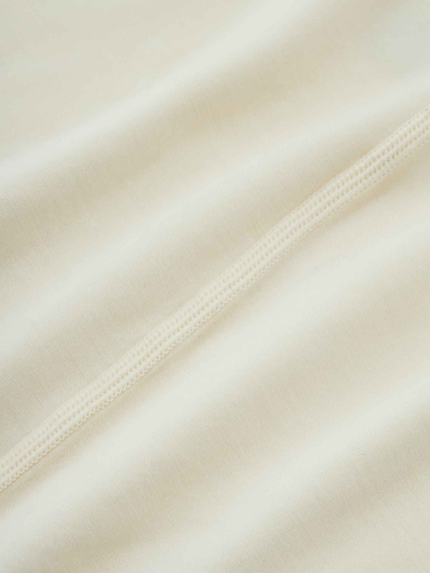 A close-up of the light cream-colored fabric of the Sno Merino Tee Men by We Norwegians reveals a smooth texture with visible stitching running diagonally across the image. The material, made from high-quality merino wool or Creora-Eco-Soft, appears soft and slightly wrinkled, emphasizing the intricate detail of the stitching.