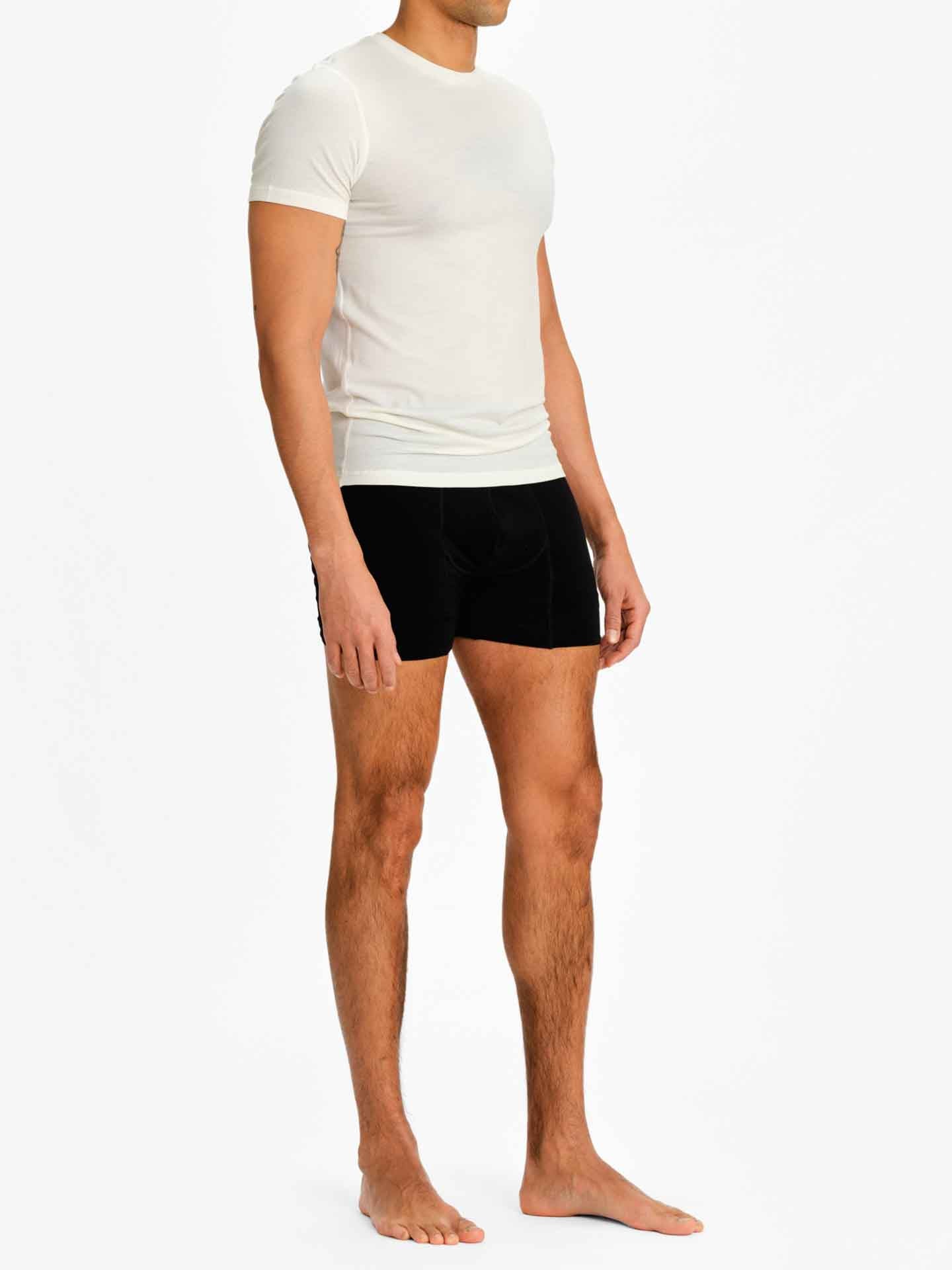 A man stands barefoot against a plain white background, donning the fitted Sno Merino Tee Men and black boxer briefs made from luxurious merino wool. Facing slightly to the side, he keeps his arms relaxed by his sides, showcasing We Norwegians' Scandinavian luxury base-layers.