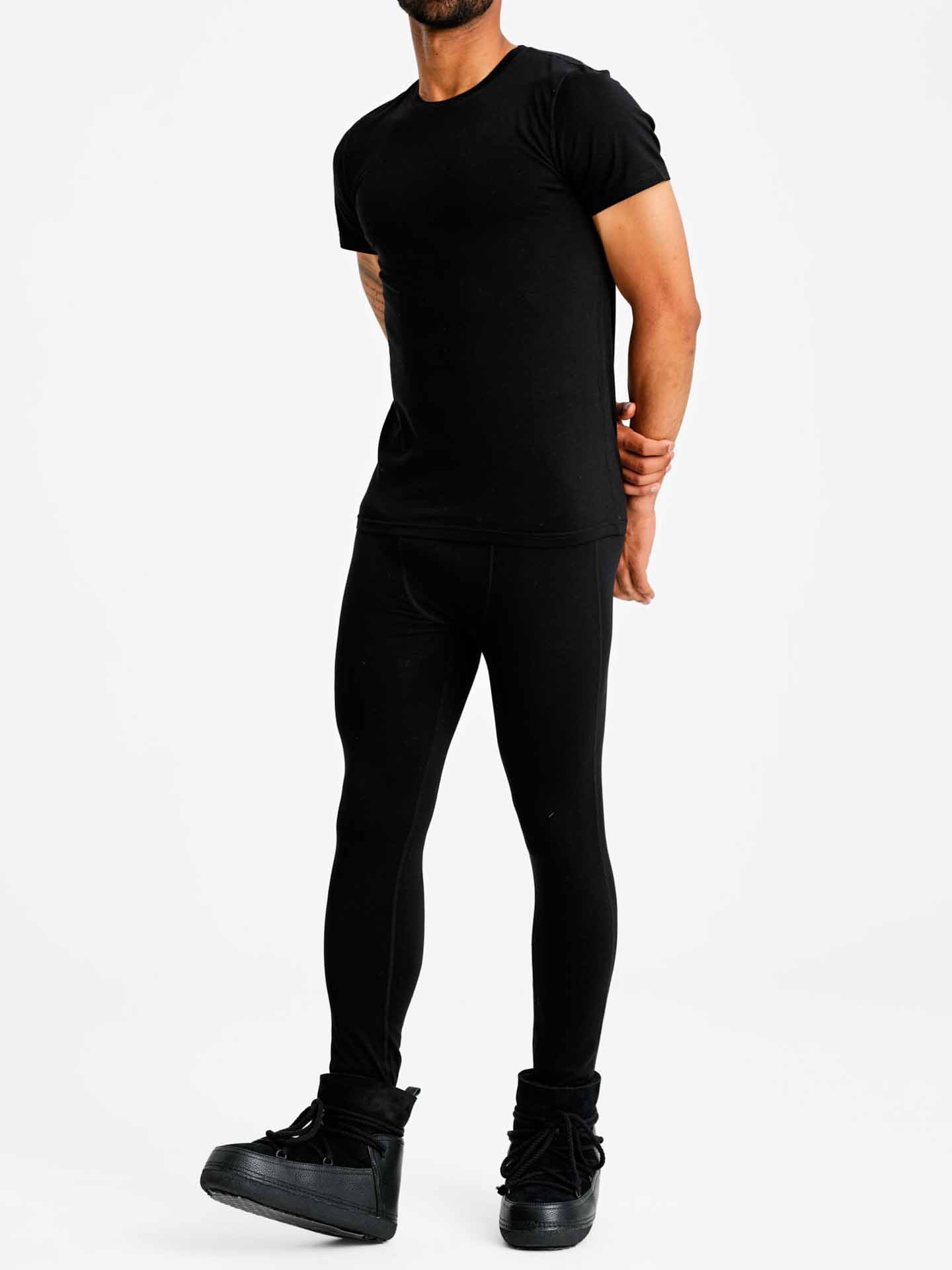 A person stands against a plain backdrop, wearing the We Norwegians Sno Merino Tee Men in black, black leggings, and black high-top sneakers. They have one hand resting on their hip and the other hanging by their side. Their face is not fully visible.