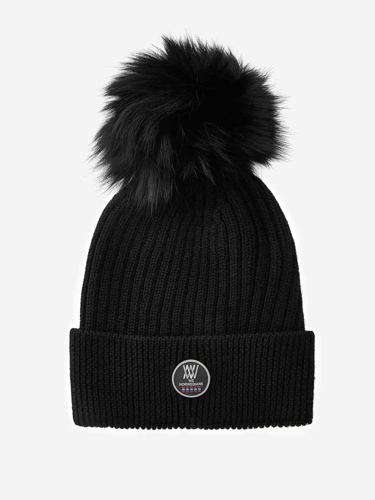 A black, ribbed knit Hemsedal Hat Women from We Norwegians features a fur pom-pom from Saga Furs. This cozy merino wool beanie includes a turn-up brim with a small round emblem showcasing a monogram logo on the front center.