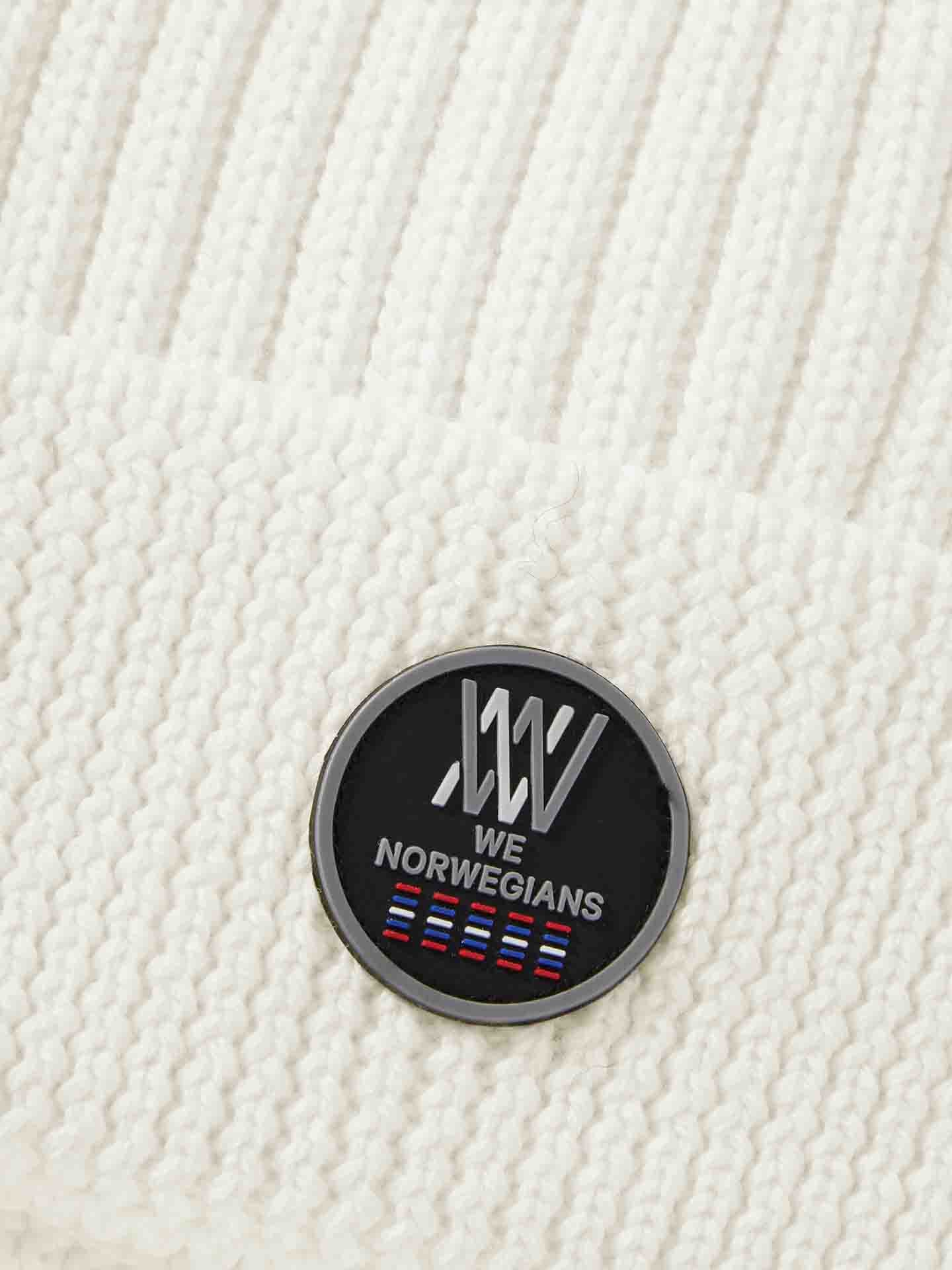 Close-up of the Hemsedal Hat Women by We Norwegians, made from white merino wool and featuring a black circular patch with the text "WE NORWEGIANS" and a logo incorporating alternating blue, red, and white stripes.