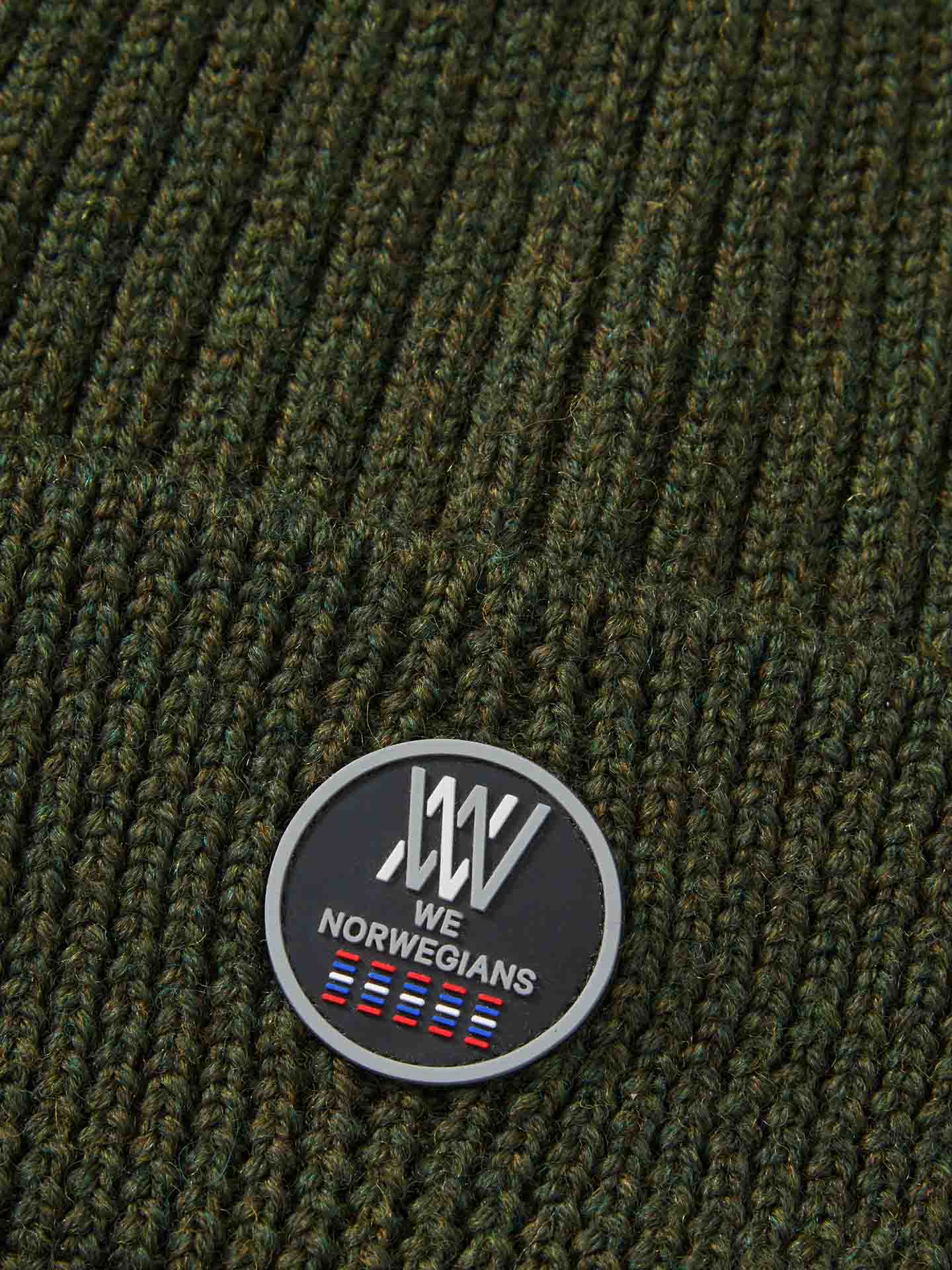 Close-up of a dark green knitted fabric, likely from the Hemsedal Hat Women by We Norwegians, featuring a circular black label stitched on it. The label displays white text "WE NORWEGIANS" along with a series of small blue and red rectangles at the bottom. Made from luxurious merino wool.