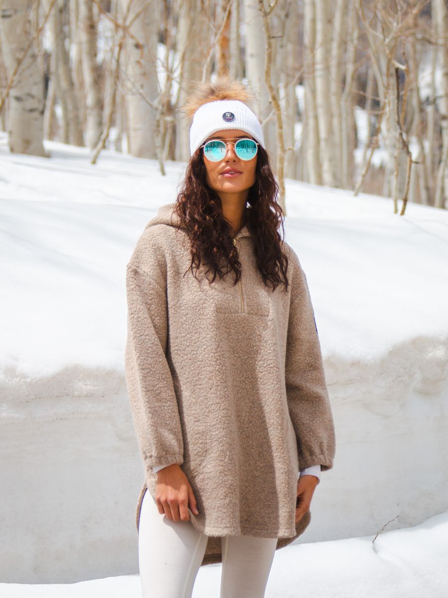 A woman stands in a snowy forest wearing a light brown fleece pullover, white We Norwegians Hemsedal Hat Women with a logo, reflective blue sunglasses, and white pants. She has curly dark hair and stands in front of snow-covered ground and bare trees.