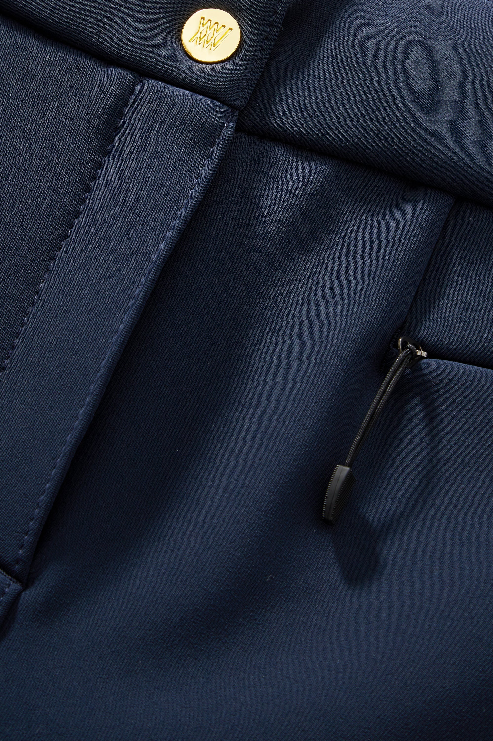 A close-up view of the Kvitfjell Bootleg Ski Pant Women by We Norwegians, showcasing fine stitching and a smooth texture. The image highlights a gold button and a small black drawstring with a zipper-like slider. These well-tailored navy blue ski pants feature brushed merino fleece and 4-way stretch soft shell for added sophistication.