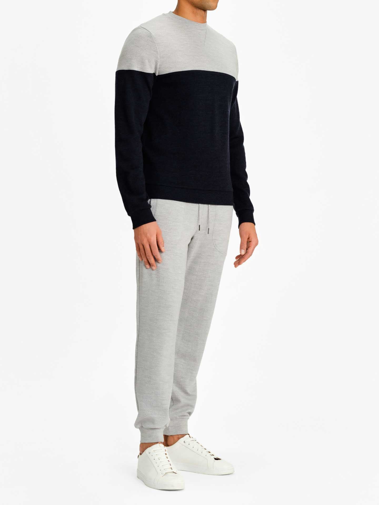 A person is dressed in a color-blocked Merino wool Tind Crewneck Men sweater by We Norwegians, featuring a light gray upper half and dark gray lower half. They are also wearing light gray Tind Joggers and white sneakers. The person is standing against a plain white background.