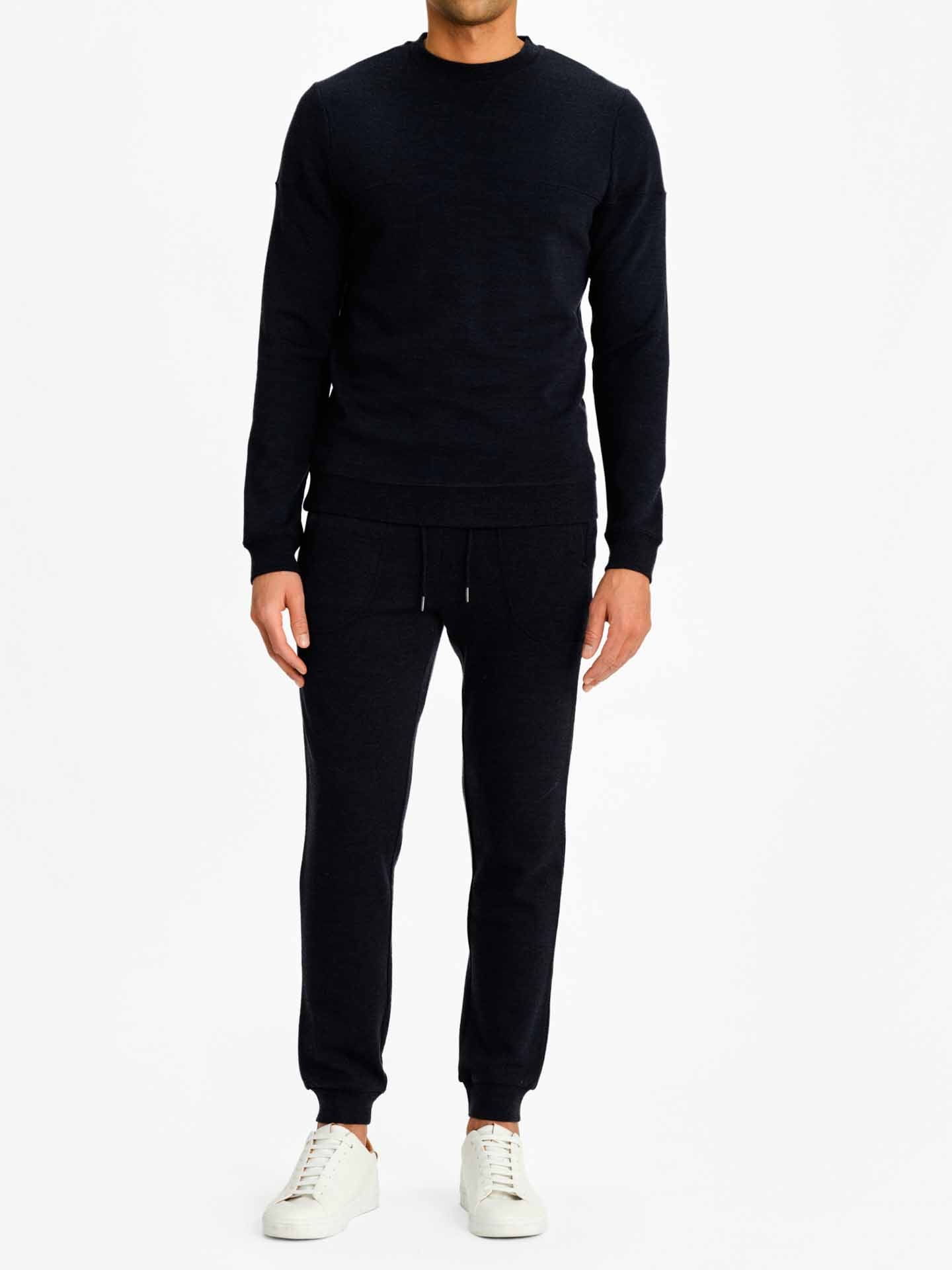 A person stands facing the camera, wearing a dark navy We Norwegians Tind Crewneck Men sweatshirt and matching Tind Joggers. They are also sporting white sneakers. The background is plain white, and the person's head is not visible in the image.