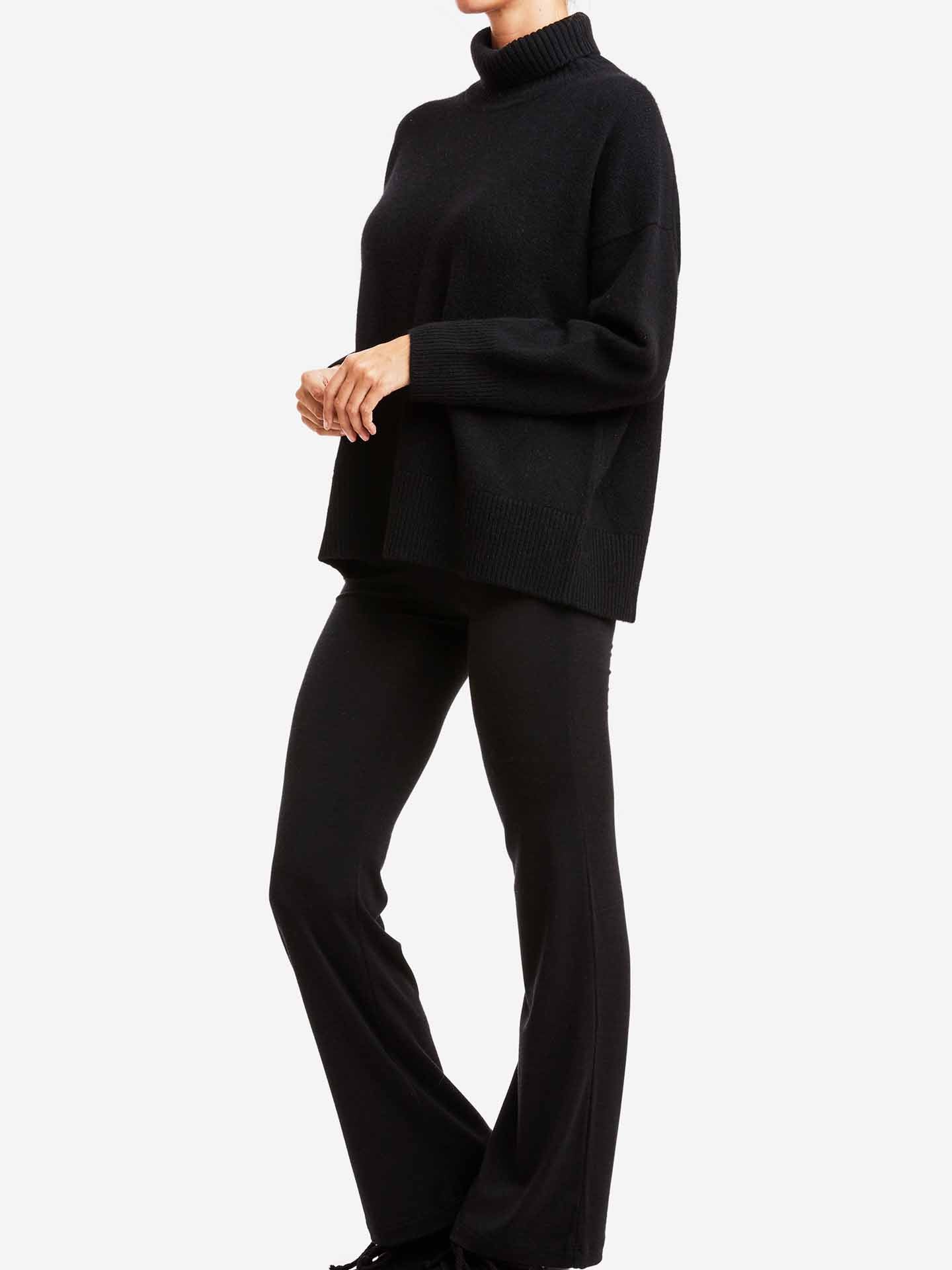 A person is standing against a plain white background, dressed in We Norwegians' Blefjell Sweater Women‚Äîa black, long-sleeved, high-neck cashmere sweater paired with black pants. The outfit appears comfortable and boasts an oversized fit. The person's head is not visible in the image.