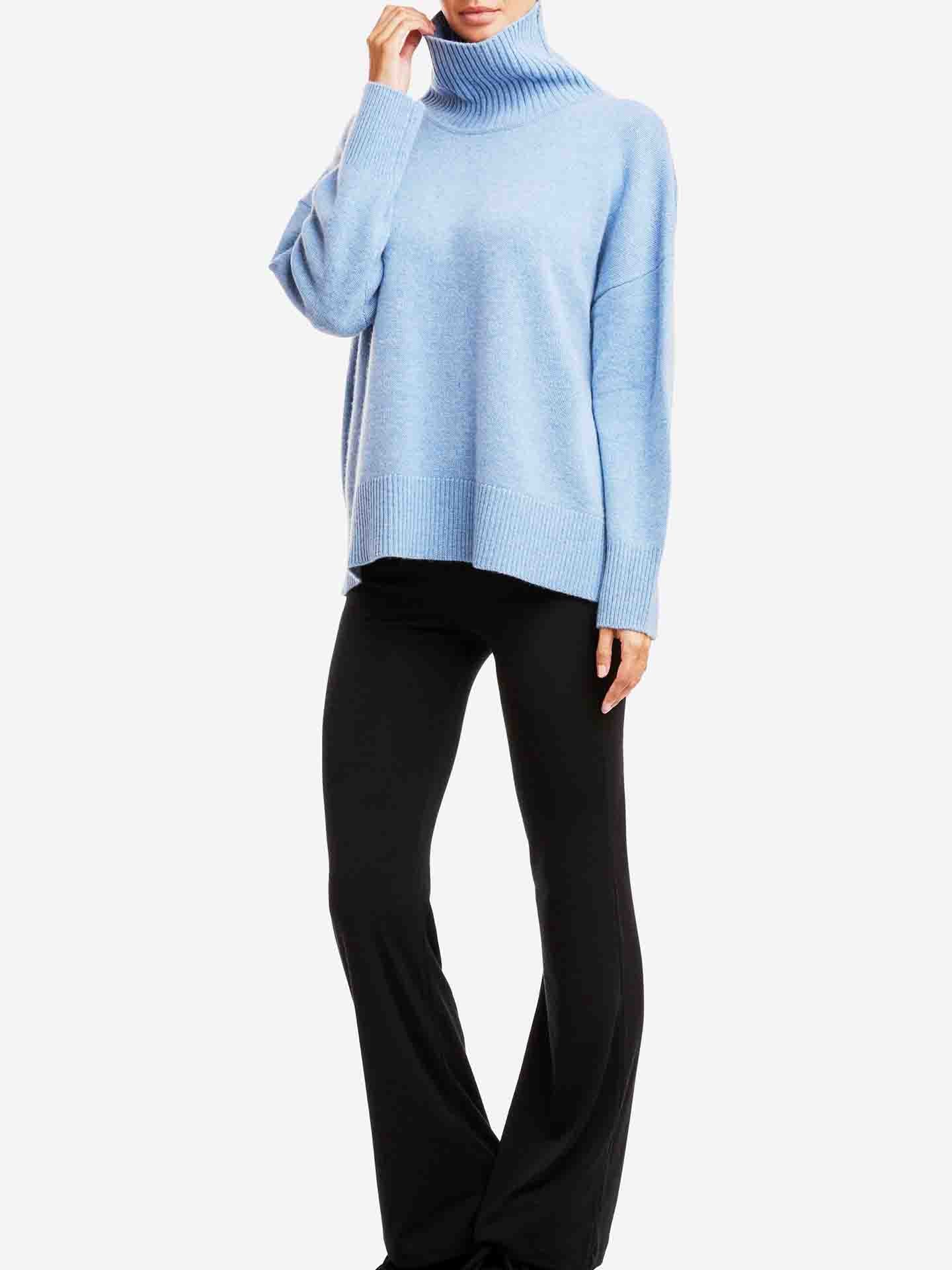 A person wearing an oversized Blefjell Sweater Women by We Norwegians in light blue cashmere, paired with black flared pants, stands against a plain white background. The person is posing with one hand touching their face and the other resting by their side.