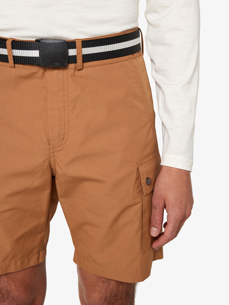 A person is wearing a white long-sleeve shirt, a We Norwegians Vidda Belt in black and white stripes, and light brown cargo shorts. Their hand is resting beside one of the shorts' pockets. The background is solid white.