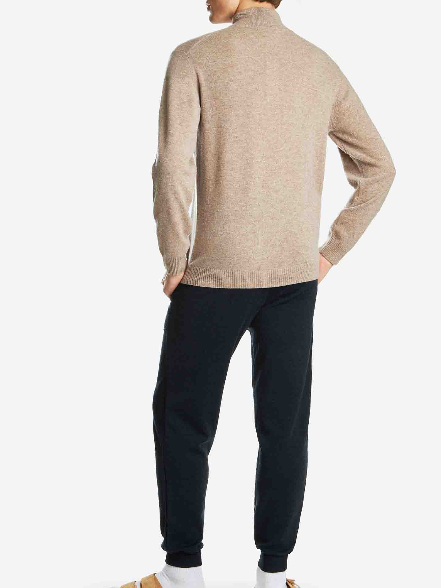 A person wearing the luxurious beige We Norwegians Blefjell Zipup Men cashmere sweater and black jogger pants is standing with their back to the camera. They are also wearing white socks and brown sandals, all set against a plain white background.