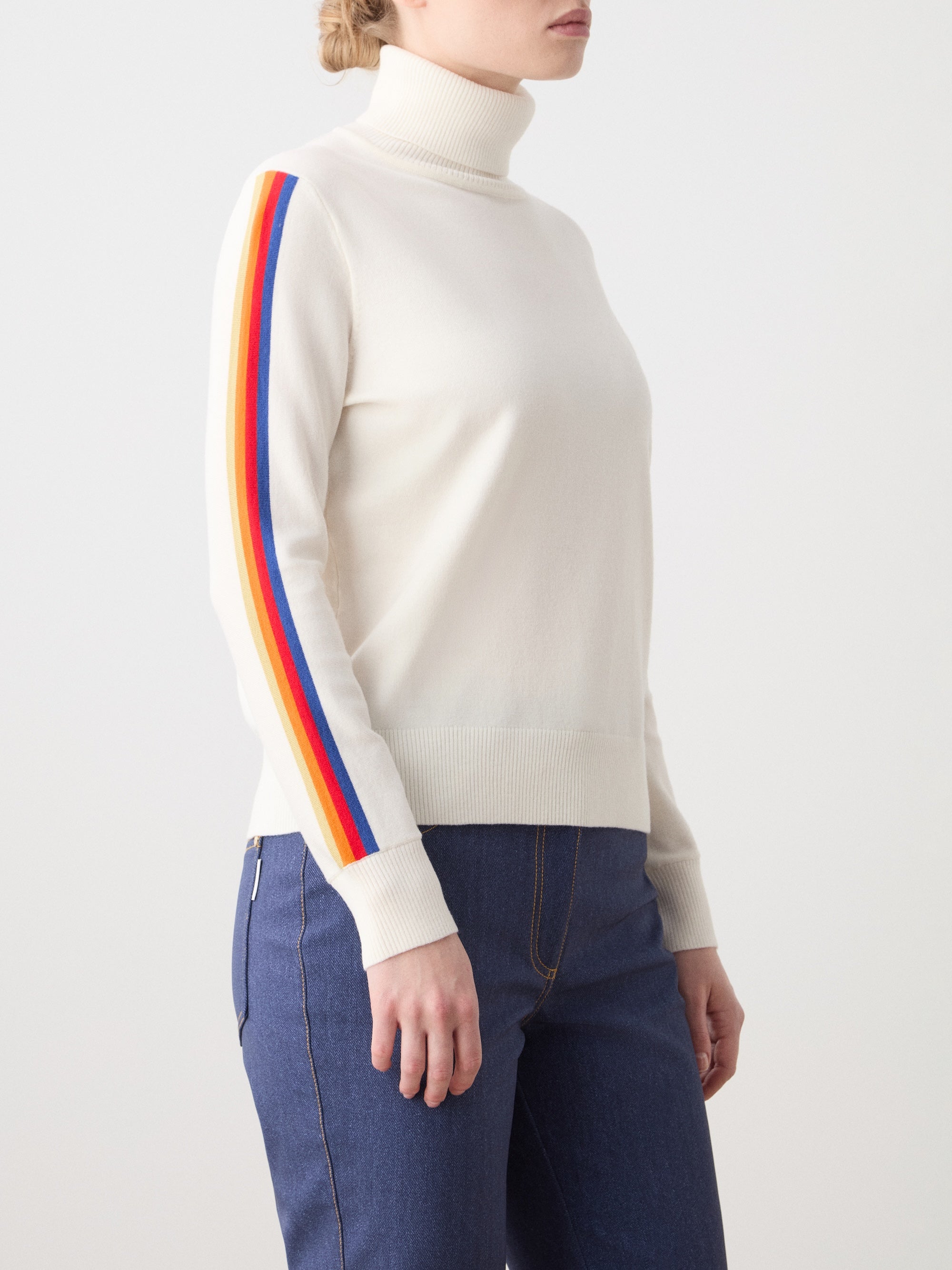 We Norwegians | FW25 | Womens Freebird Turtleneck Sweater_Off-white | Model is wearing this knitted turtleneck sweater with contrasting panels on the sleeves - paired with the Western ski pants | Shot against an off-white background | Side/ Close-up