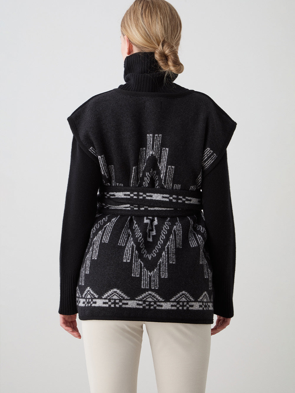 We Norwegians_FW25_Women | Model is wearing the Kautokeino vest | Black in color with a jacquard pattern inspired by Native and Indigenous American Art | Vest has a matching belt to cinch the waist | Back view on a white background