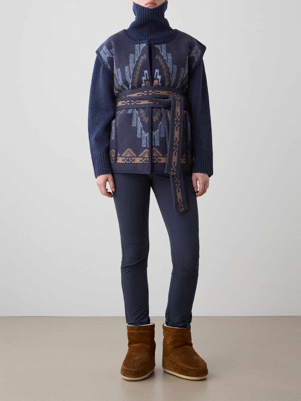 We Norwegians_FW25_Women | Model is wearing the Kautokeino vest | Navy blue in color with a jacquard pattern inspired by Native and Indigenous American Art | Vest has a matching belt to cinch the waist | Front view on a white background