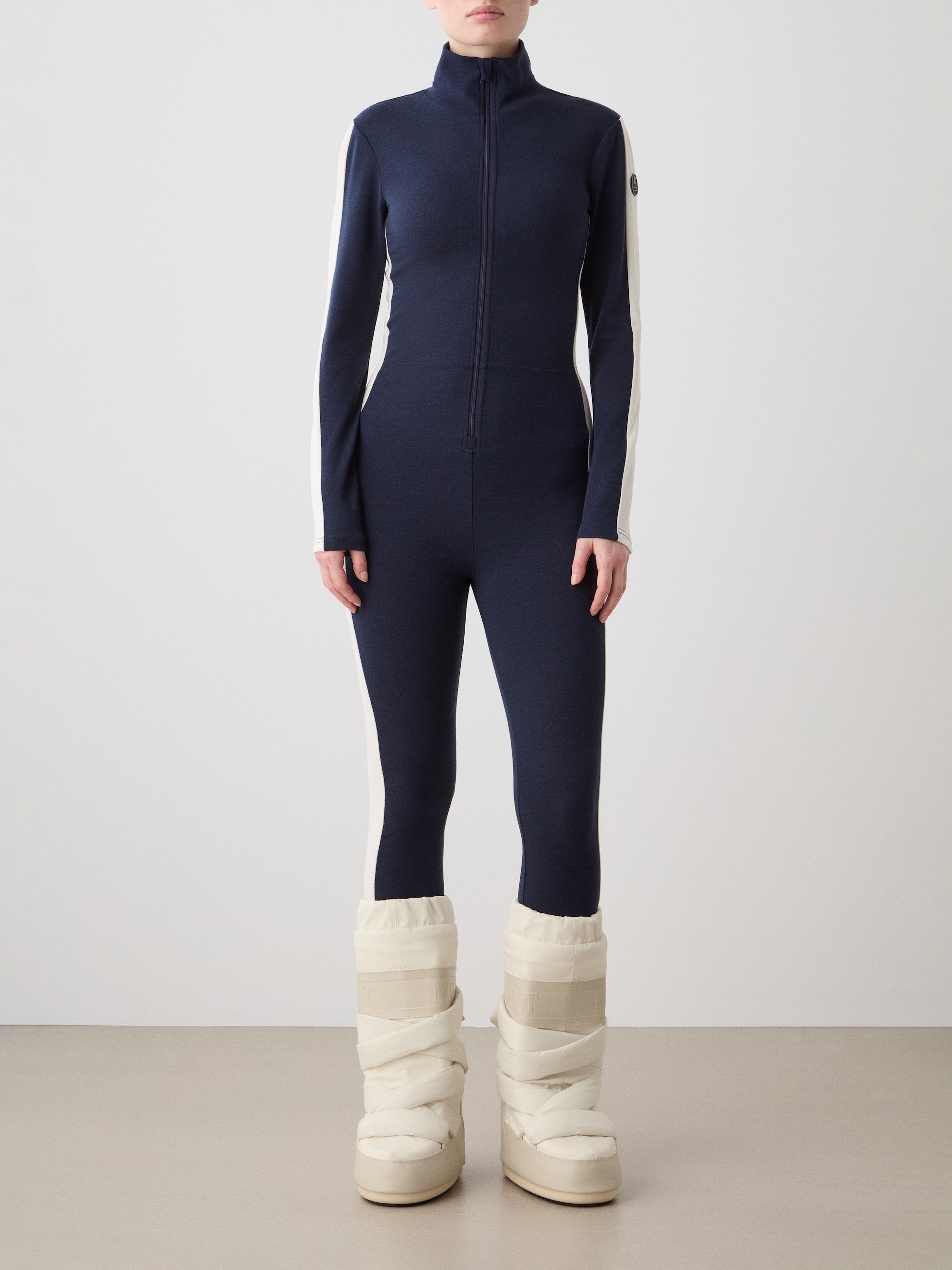 We Norwegians | FW25 | Womens Voss Jumpsuit_Navy blue | The model is wearing this piqué knit jumpsuit with a front zipper opening, a stand collar, white panels on the sleeves with subtle branding - paired with white snow boots | Shot against an off-white background | Front