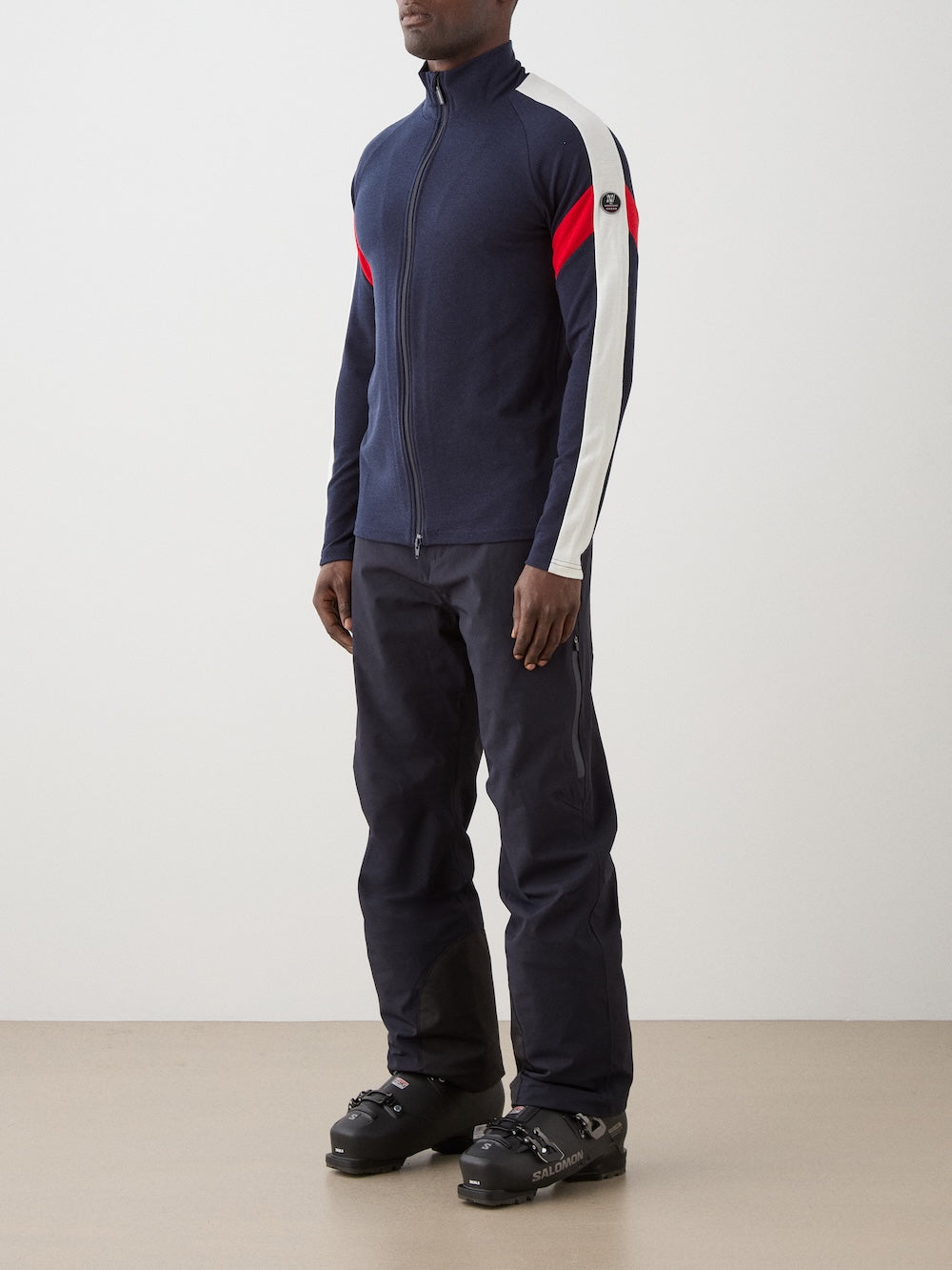 We Norwegians | FW25 | Mens Zermatt Full Zip Sweater_Flag | The model is wearing this piqué knit sweater with white and red accents on the sleeves and a front 2-way zipper opening - paired with black ski pants | Shot against an off-white background | Side