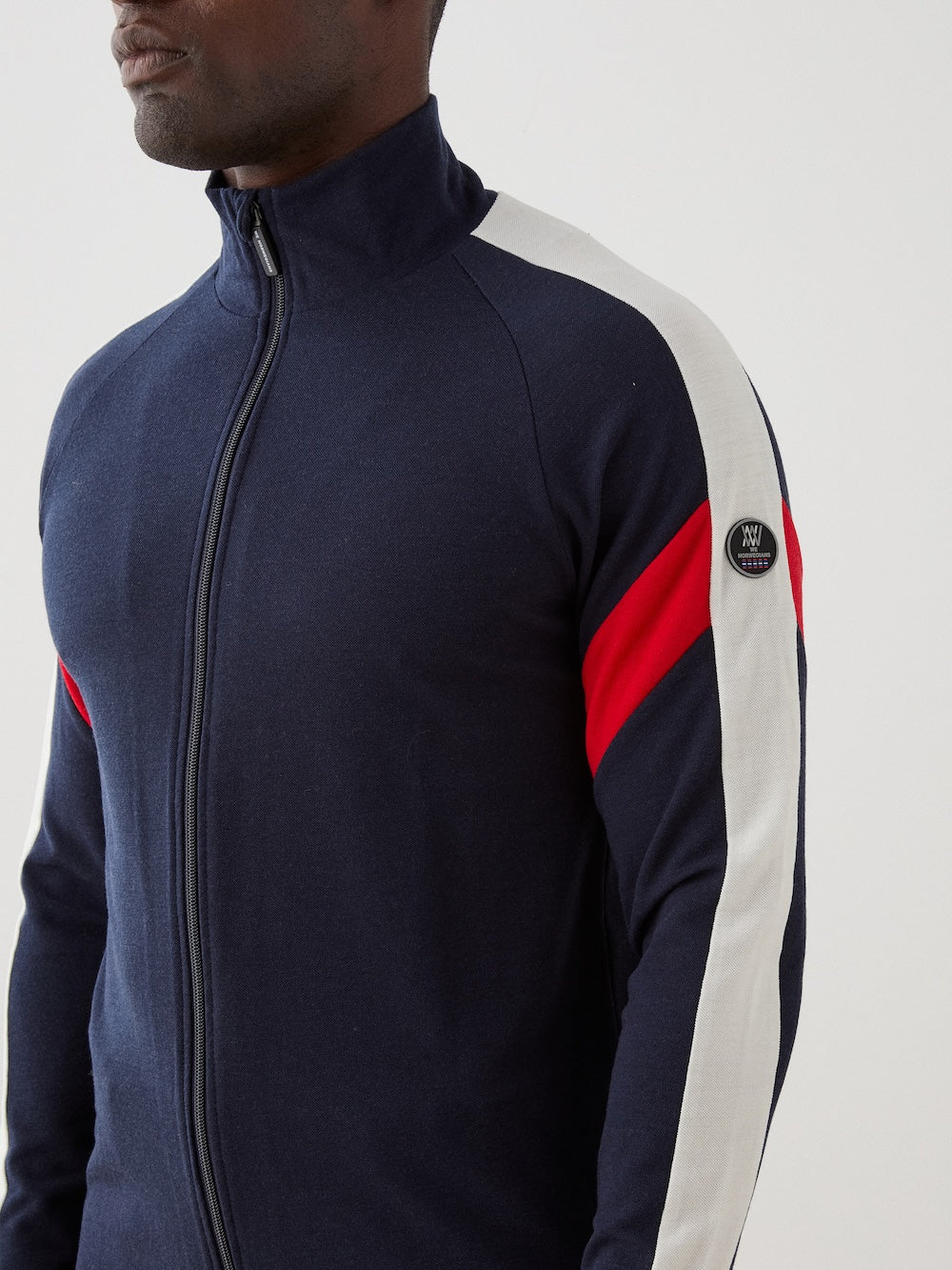 We Norwegians | FW25 | Mens Zermatt Full Zip Sweater_Flag | The model is wearing this piqué knit sweater with white and red accents on the sleeves and a front 2-way zipper opening | Shot against an off-white background | Close-up