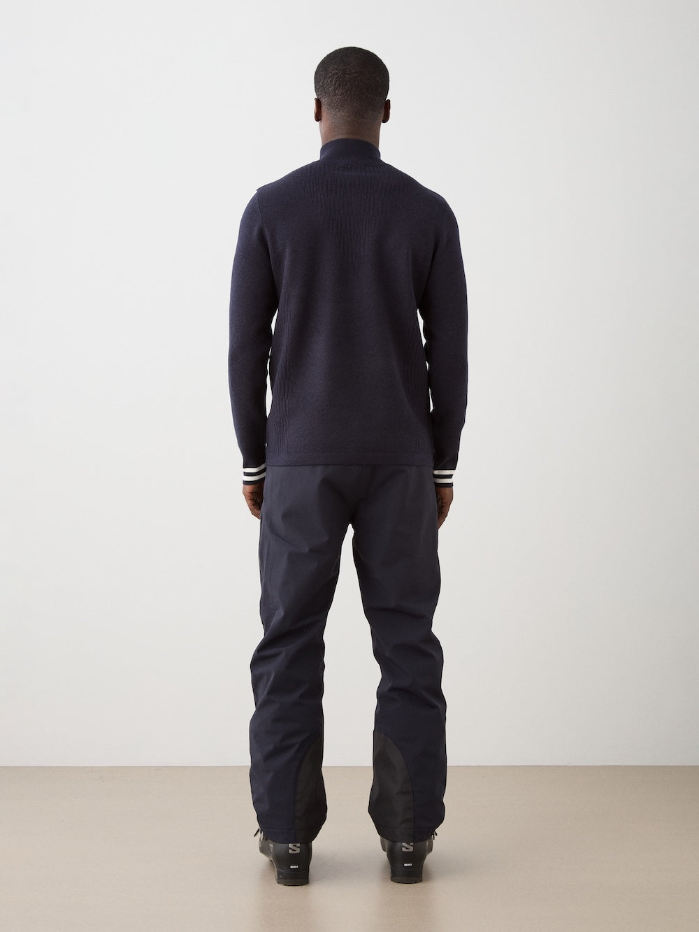 We Norwegians | FW25 | Mens Rocky ZipUp_Navy blue | The model is wearing this knitted sweater designed in a retro style with a front 1/4th zipper opening, color block panels on the sleeves, with embroidered branding - paired with black ski pants | Back